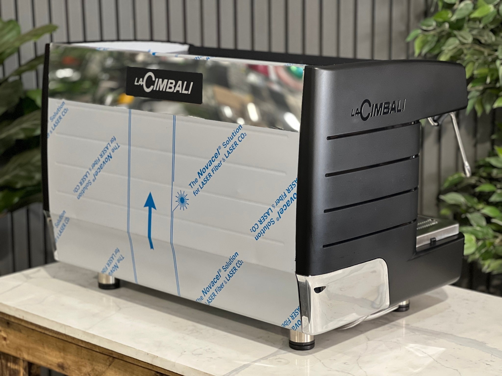 A brand new La Cimbali M23 UP 2 Group Coffee Machine with Black finish sitting on a marble bench. There is greenery in the background. The image is taken from behind the machine and the machine is sitting on an angle so that you can see the back of the machine and the side.