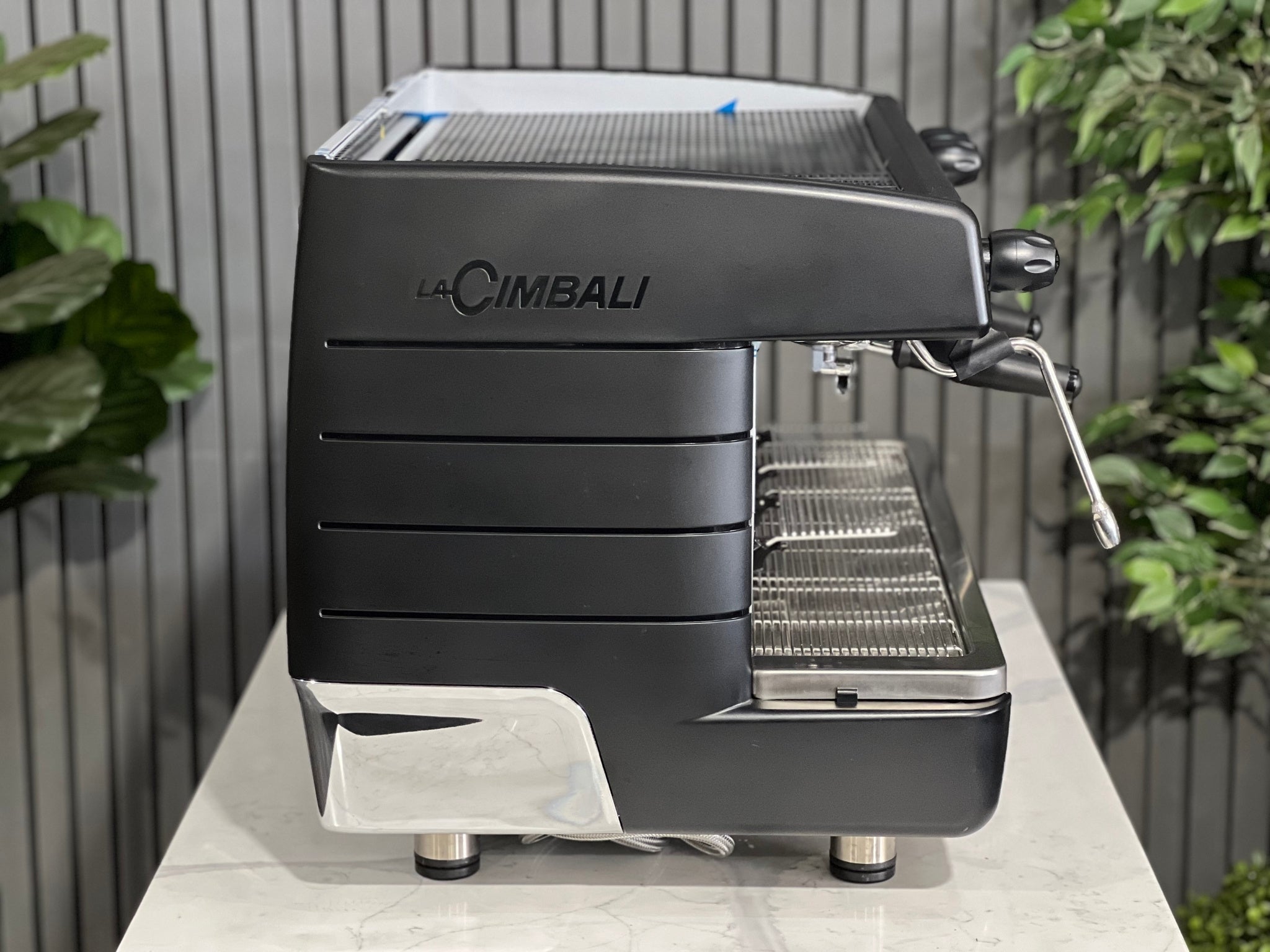 A brand new La Cimbali M23 UP 2 Group Coffee Machine with Black finish sitting on a marble bench. There is greenery in the background. The image is taken from the side of the machine.
