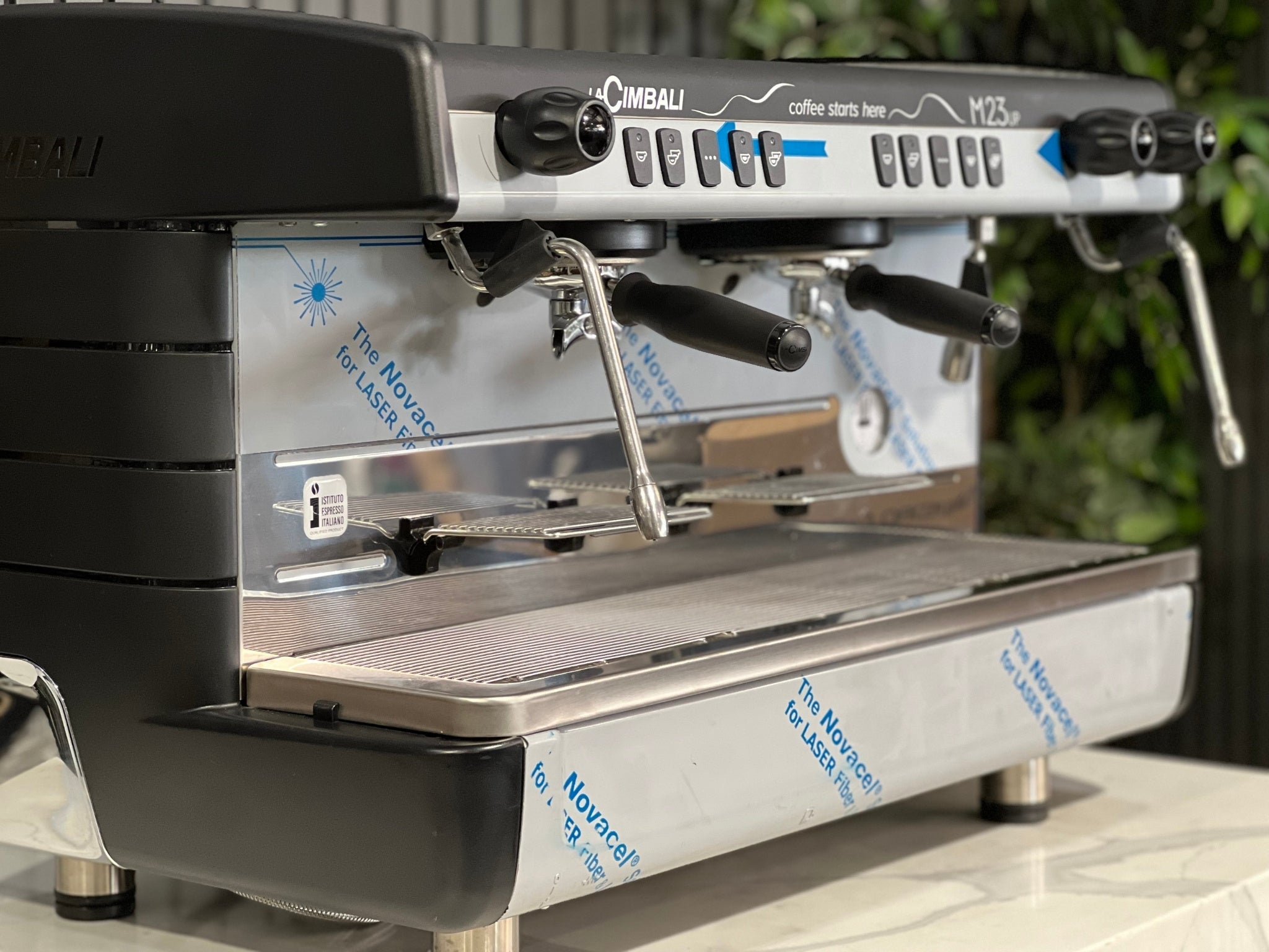 A brand new La Cimbali M23 UP 2 Group Coffee Machine with Black finish s sitting on a marble bench. There is greenery in the background. The image is a close up image taken from the front of the machine and the machine is sitting on an angle so that you can see the front of the machine, the controls including buttons, group handles and logo.