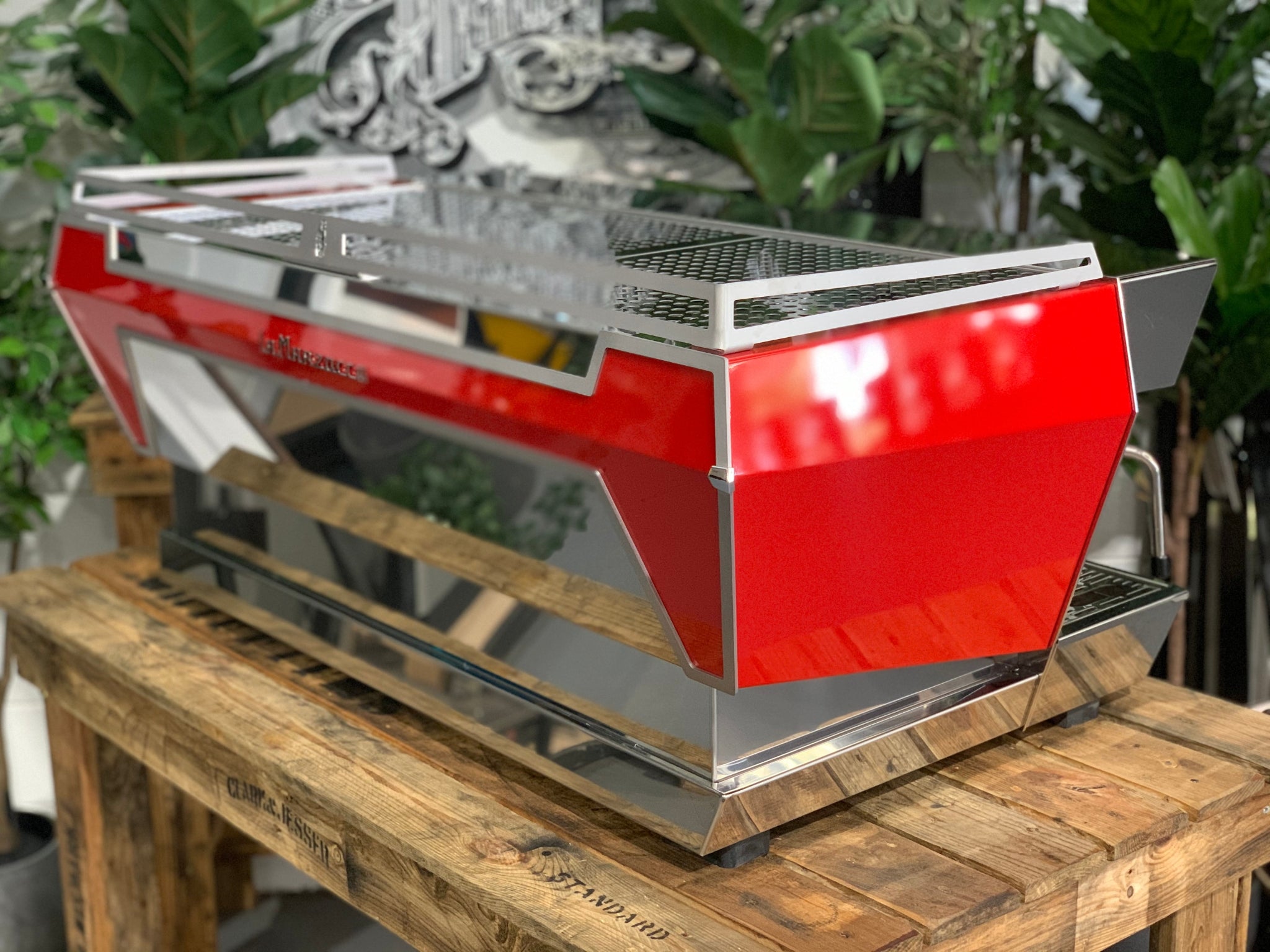 Angled rear view of La Marzocco KB90 3 Group Red & Stainless Coffee Machine - New showing side and back panels and stainless steel finish on a wooden bench with greenery, espresso machine.