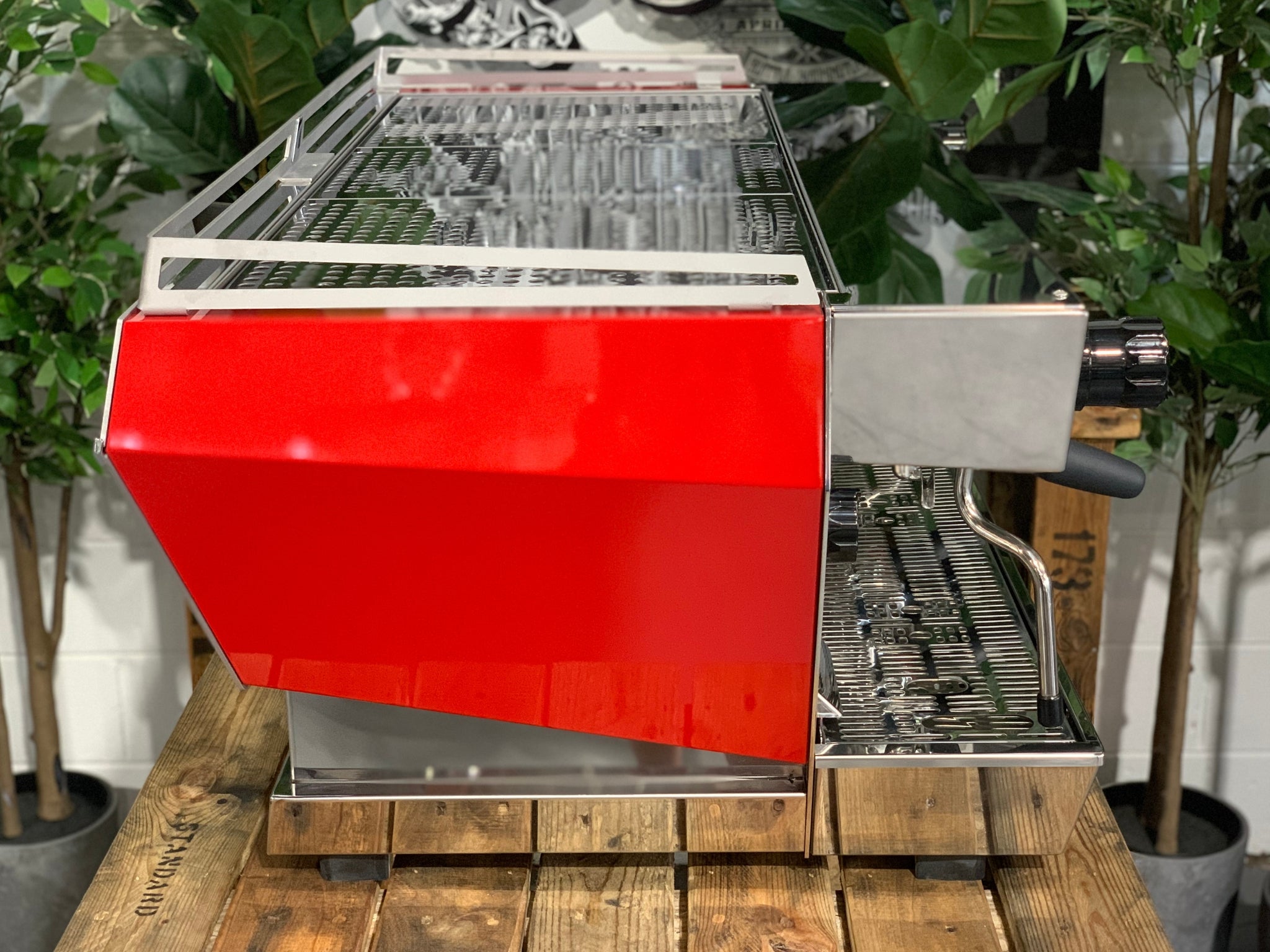 Side view of La Marzocco KB90 3 Group Red & Stainless Coffee Machine - New showing red and stainless steel body and ergonomic portafilters on a wooden bench with greenery, espresso coffee machine.