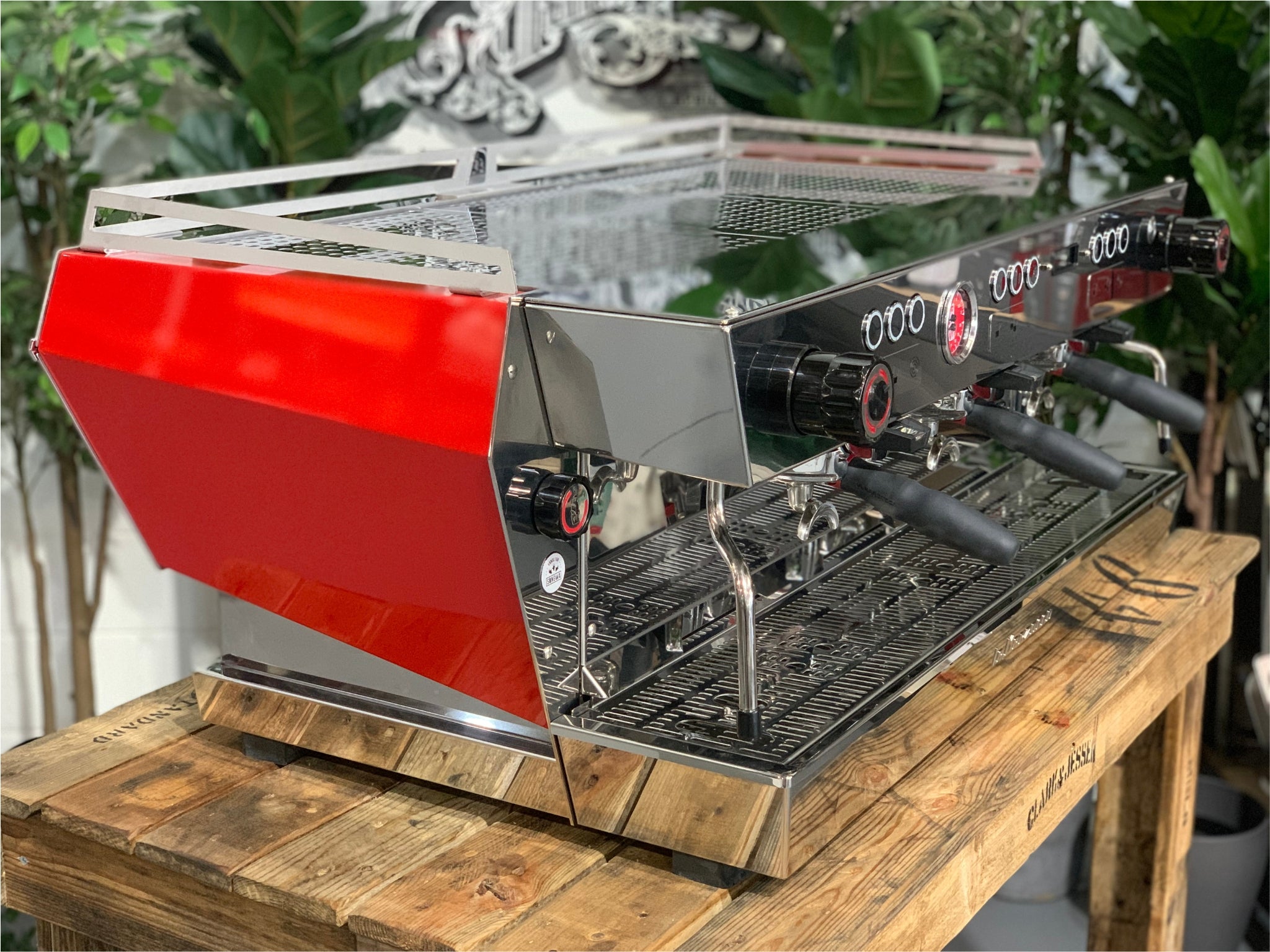 La Marzocco KB90 3 Group Red & Stainless Coffee Machine - New angled view showing three group heads, ergonomic straight-in portafilters, and stainless steel body on a wooden bench with greenery in the background, espresso coffee machine.