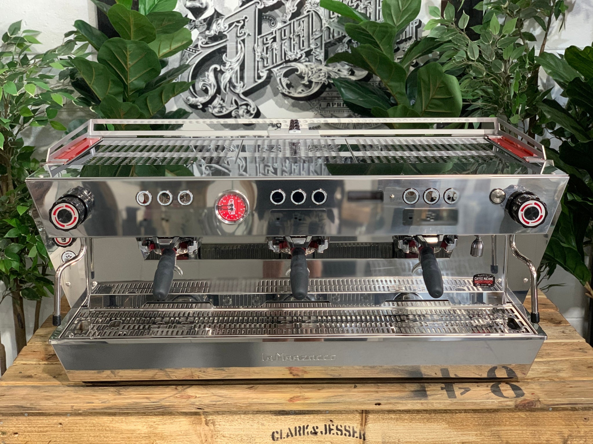 Top-front view of La Marzocco KB90 3 Group Red & Stainless Coffee Machine - New showing control buttons, three group heads, and ergonomic portafilters on a wooden bench with greenery, espresso coffee machine.