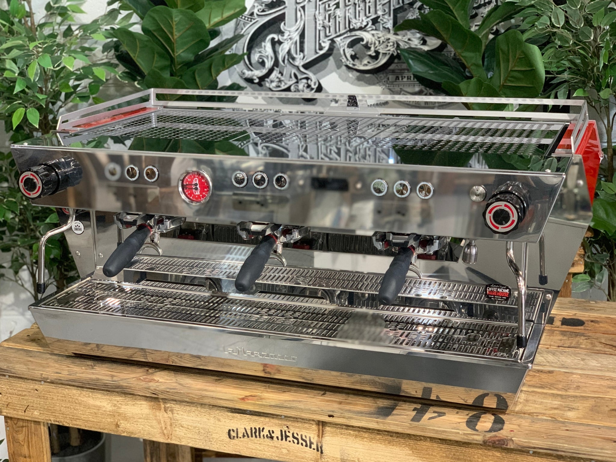 La Marzocco KB90 3 Group Red & Stainless Coffee Machine - New angled view showing three group heads, ergonomic straight-in portafilters, and stainless steel body on a wooden bench with greenery in the background, espresso coffee machine.
