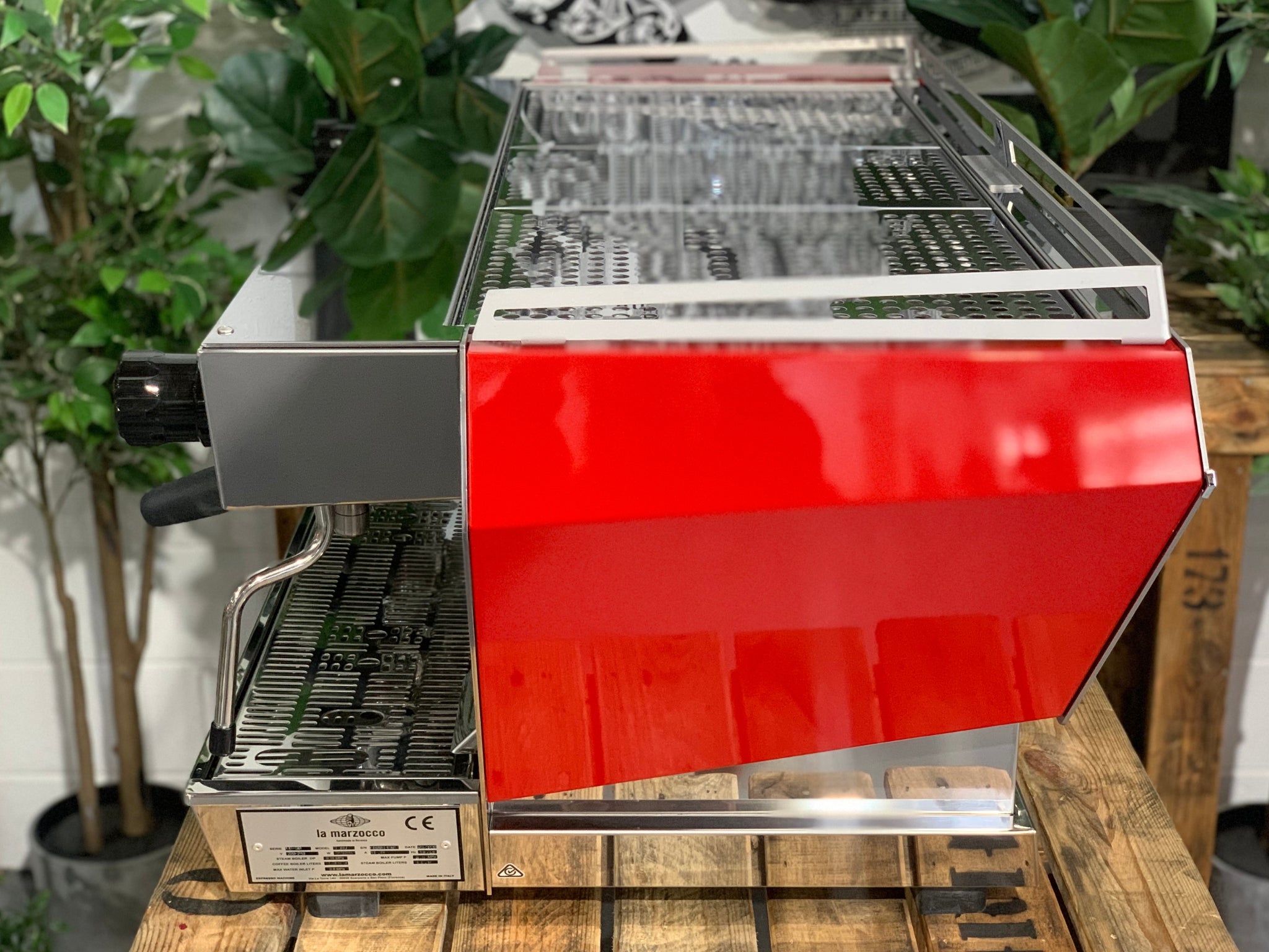 Side view of La Marzocco KB90 3 Group Red & Stainless Coffee Machine - New showing red and stainless steel body and ergonomic portafilters on a wooden bench with greenery, espresso coffee machine.