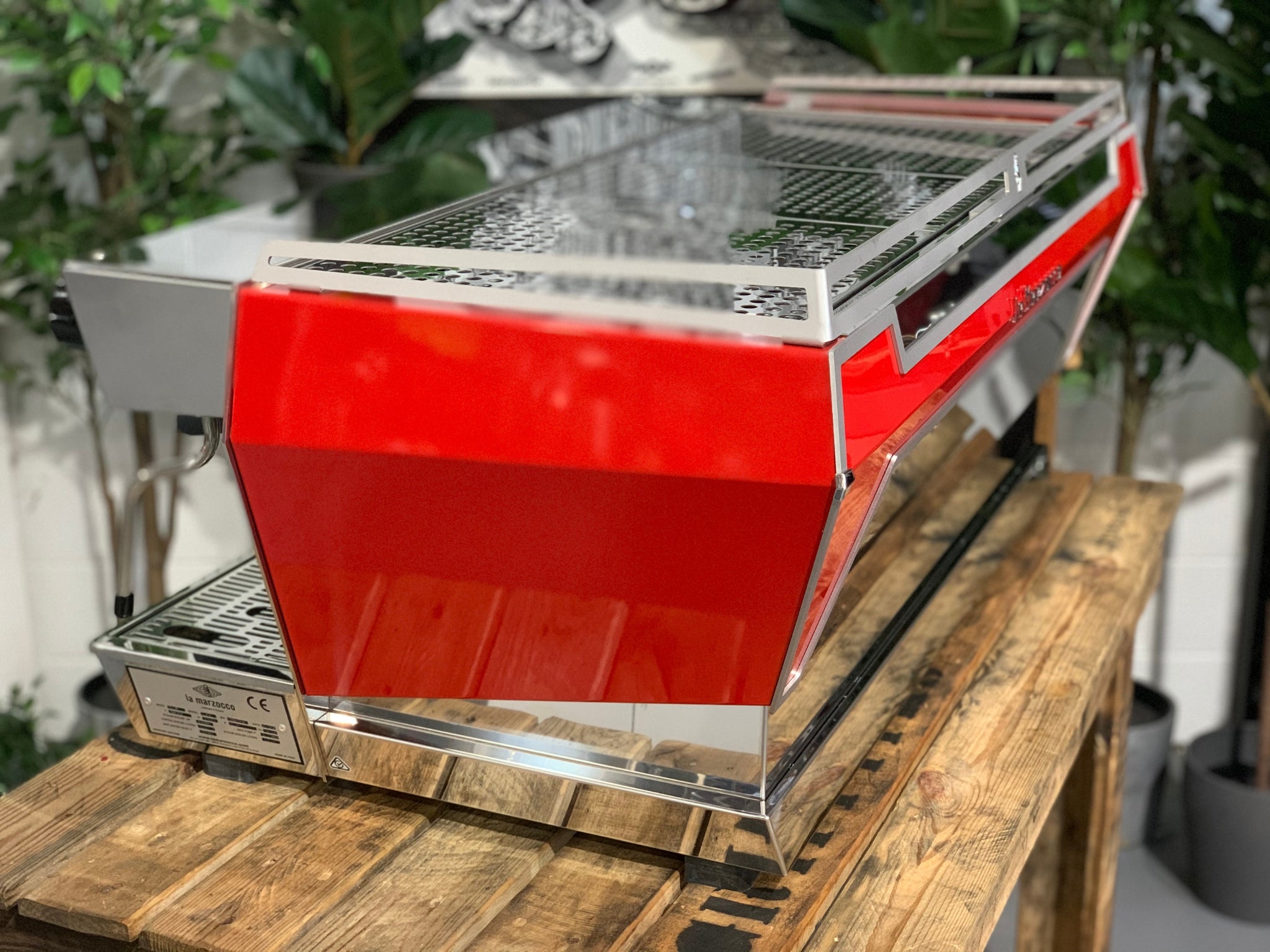 Angled rear view of La Marzocco KB90 3 Group Red & Stainless Coffee Machine - New showing side and back panels and stainless steel finish on a wooden bench with greenery, espresso machine.