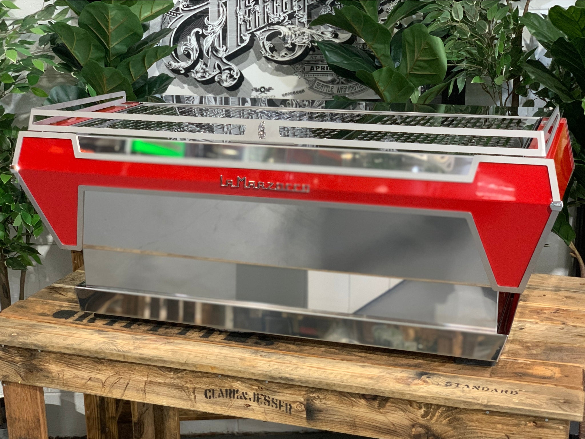 Angled rear view of La Marzocco KB90 3 Group Red & Stainless Coffee Machine - New showing side and back panels and stainless steel finish on a wooden bench with greenery, espresso machine.