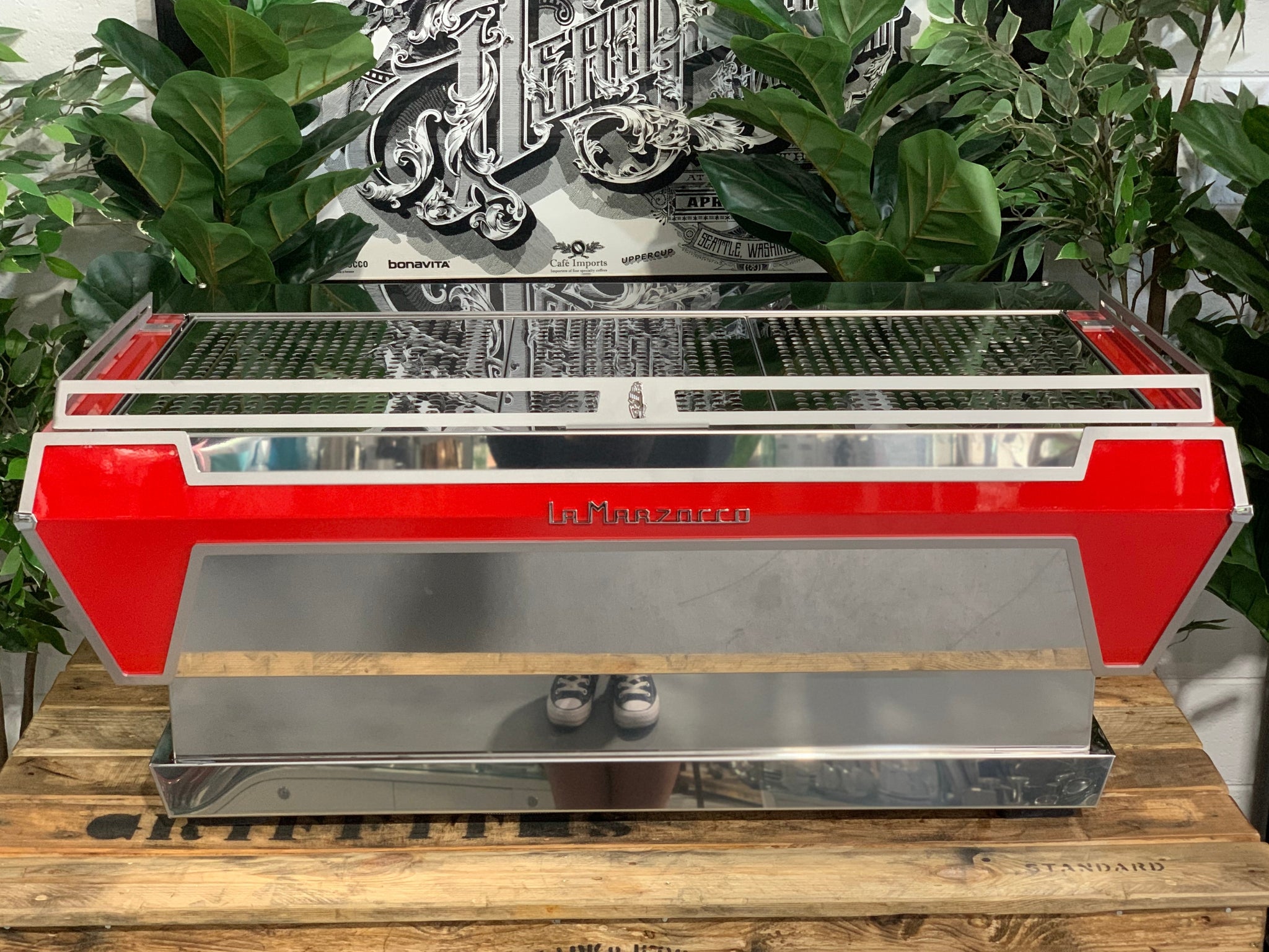 Rear view of La Marzocco KB90 3 Group Red & Stainless Coffee Machine - New showing stainless steel back panel and overall machine profile on a wooden bench with greenery, espresso machine.