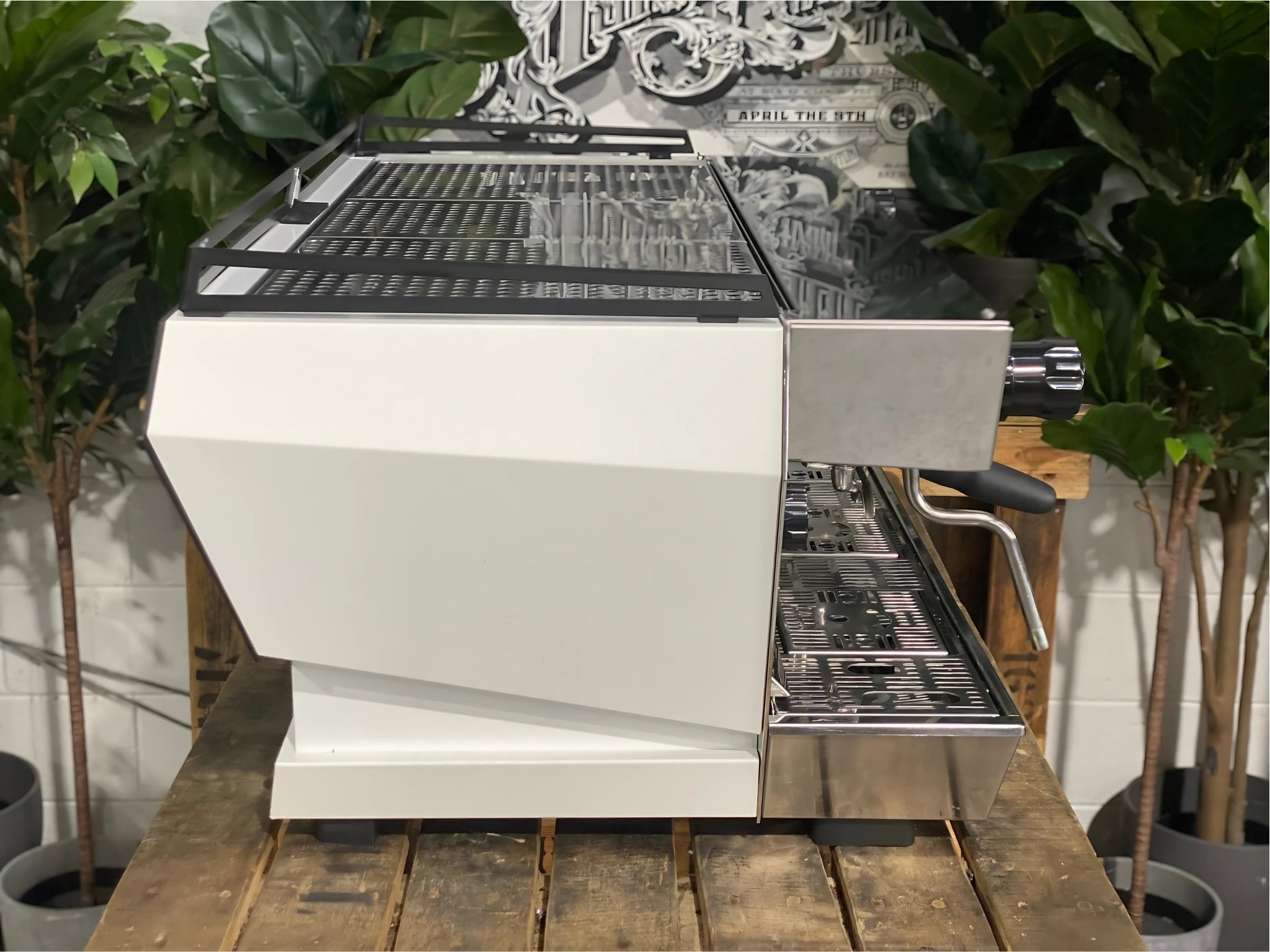 La Marzocco KB90 ABR 3 Group Coffee Machine White side view revealing the smooth side panel, cool touch steam wand, and part of the group head, designed for heavy-duty espresso performance on a wooden bench with greenery in the background.