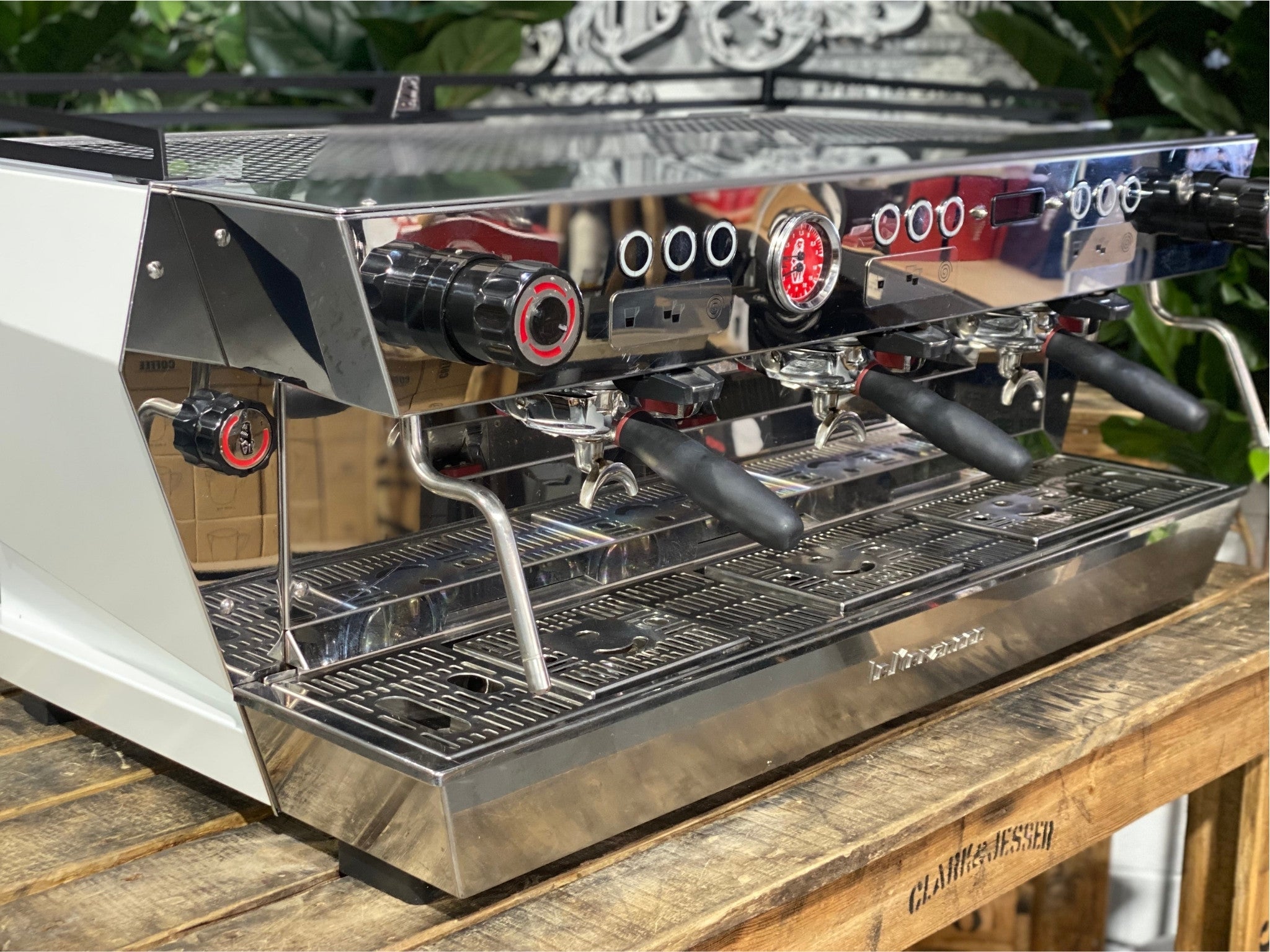 La Marzocco KB90 ABR 3 Group Coffee Machine White close-up front detail showing the group head, straight-in portafilter, and integrated scale for precision espresso brewing, with wooden bench and greenery providing a natural setting.