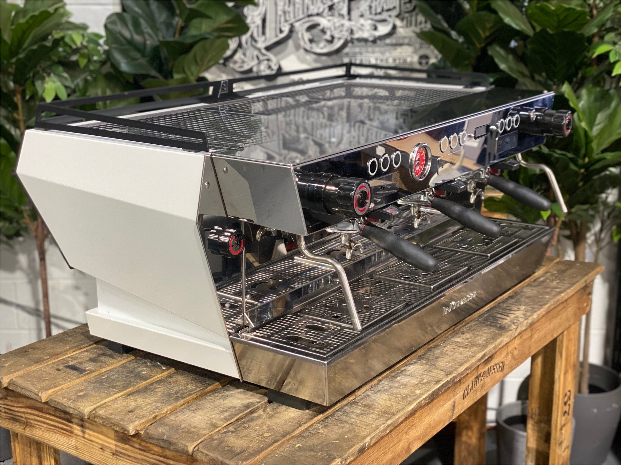 La Marzocco KB90 ABR 3 Group Coffee Machine White angled view showing the sleek white body, triple group heads, and cool touch steam wands of this high-performance commercial espresso machine, sitting on a wooden bench with greenery in the background.