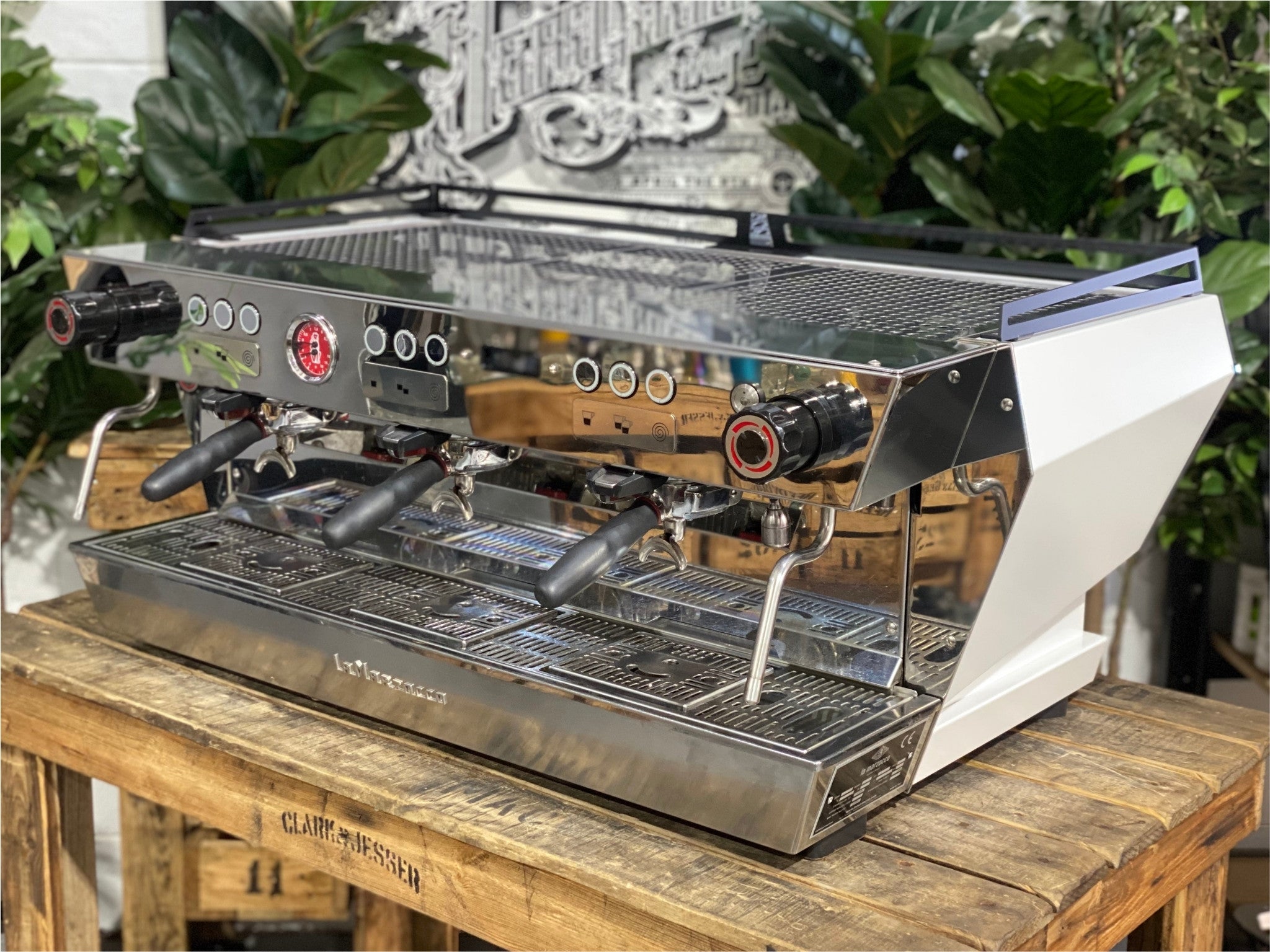 La Marzocco KB90 ABR 3 Group Coffee Machine White angled view showing the sleek white body, triple group heads, and cool touch steam wands of this high-performance commercial espresso machine, sitting on a wooden bench with greenery in the background.