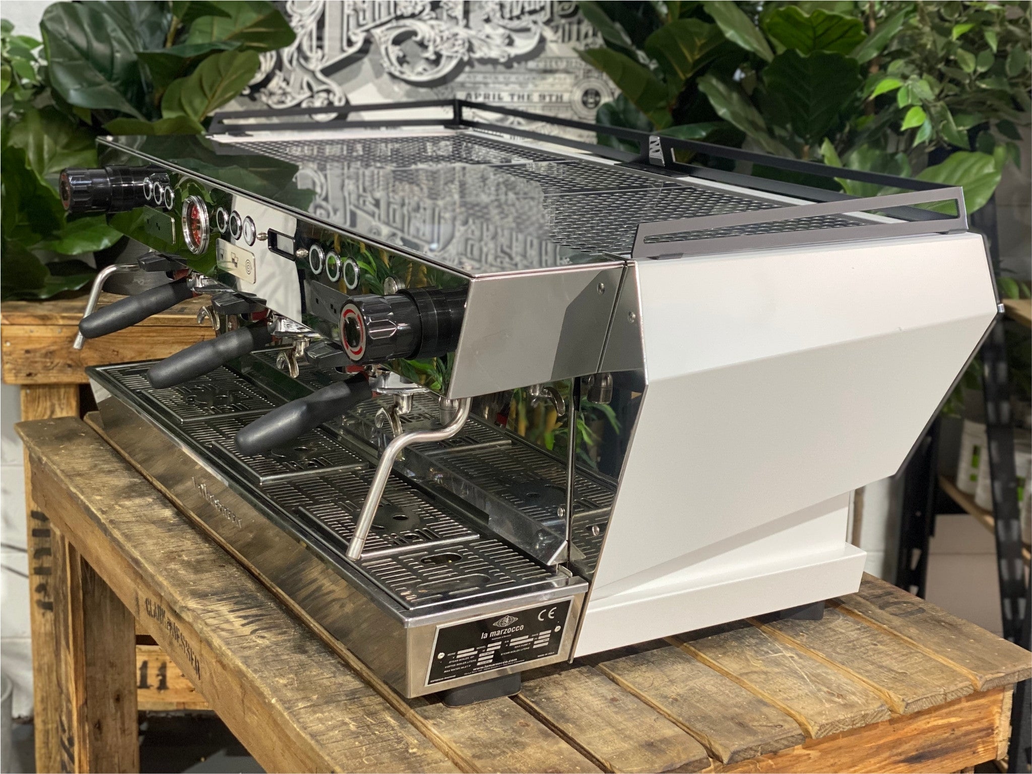 La Marzocco KB90 ABR 3 Group Coffee Machine White side view revealing the smooth side panel, cool touch steam wand, and part of the group head, designed for heavy-duty espresso performance on a wooden bench with greenery in the background.