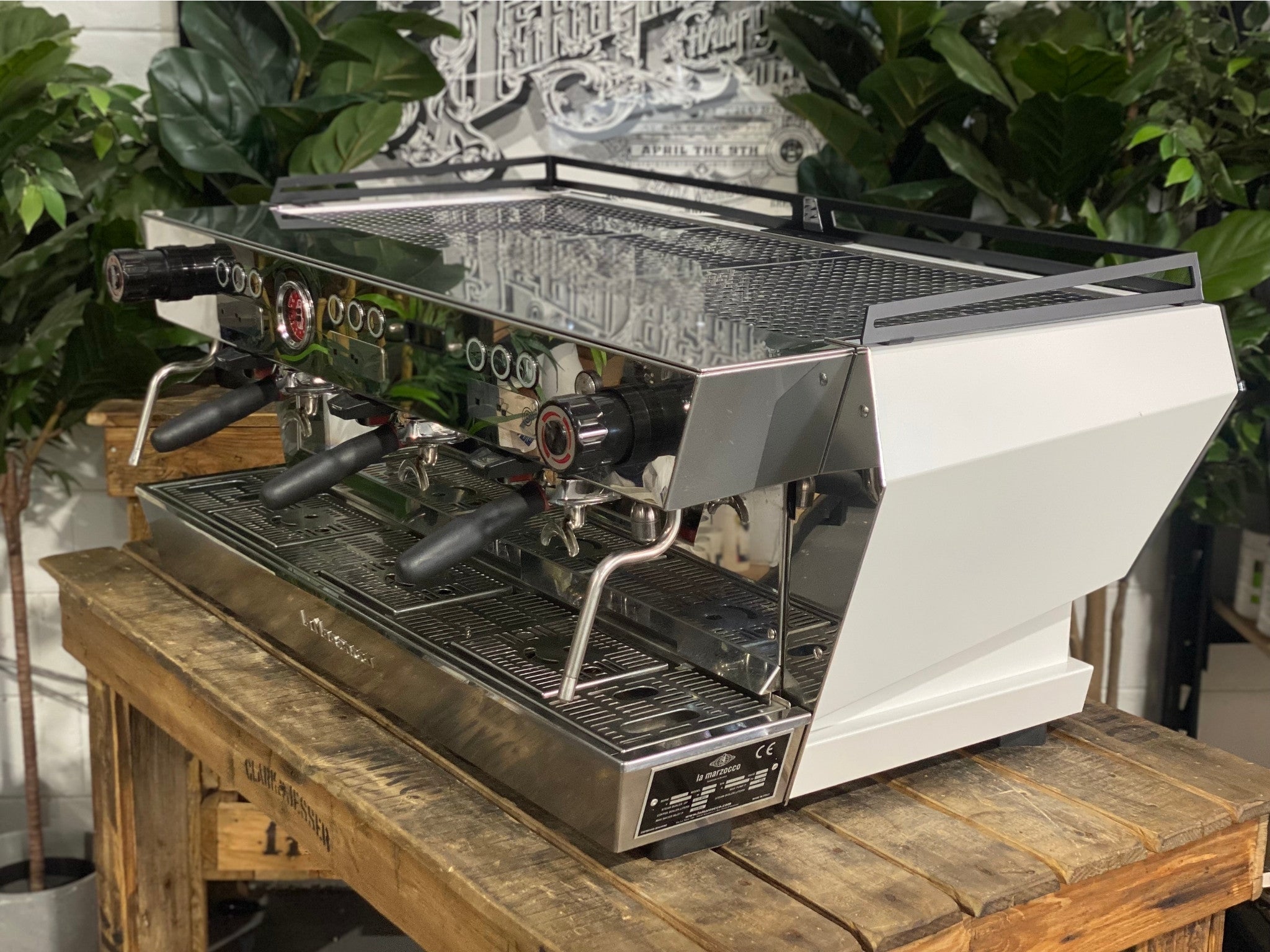La Marzocco KB90 ABR 3 Group Coffee Machine White side view revealing the smooth side panel, cool touch steam wand, and part of the group head, designed for heavy-duty espresso performance on a wooden bench with greenery in the background.