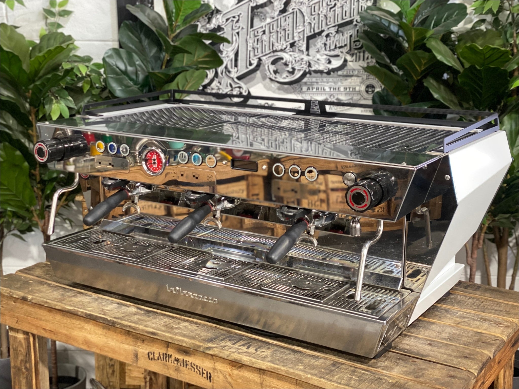 La Marzocco KB90 ABR 3 Group Coffee Machine White angled view showing the sleek white body, triple group heads, and cool touch steam wands of this high-performance commercial espresso machine, sitting on a wooden bench with greenery in the background.