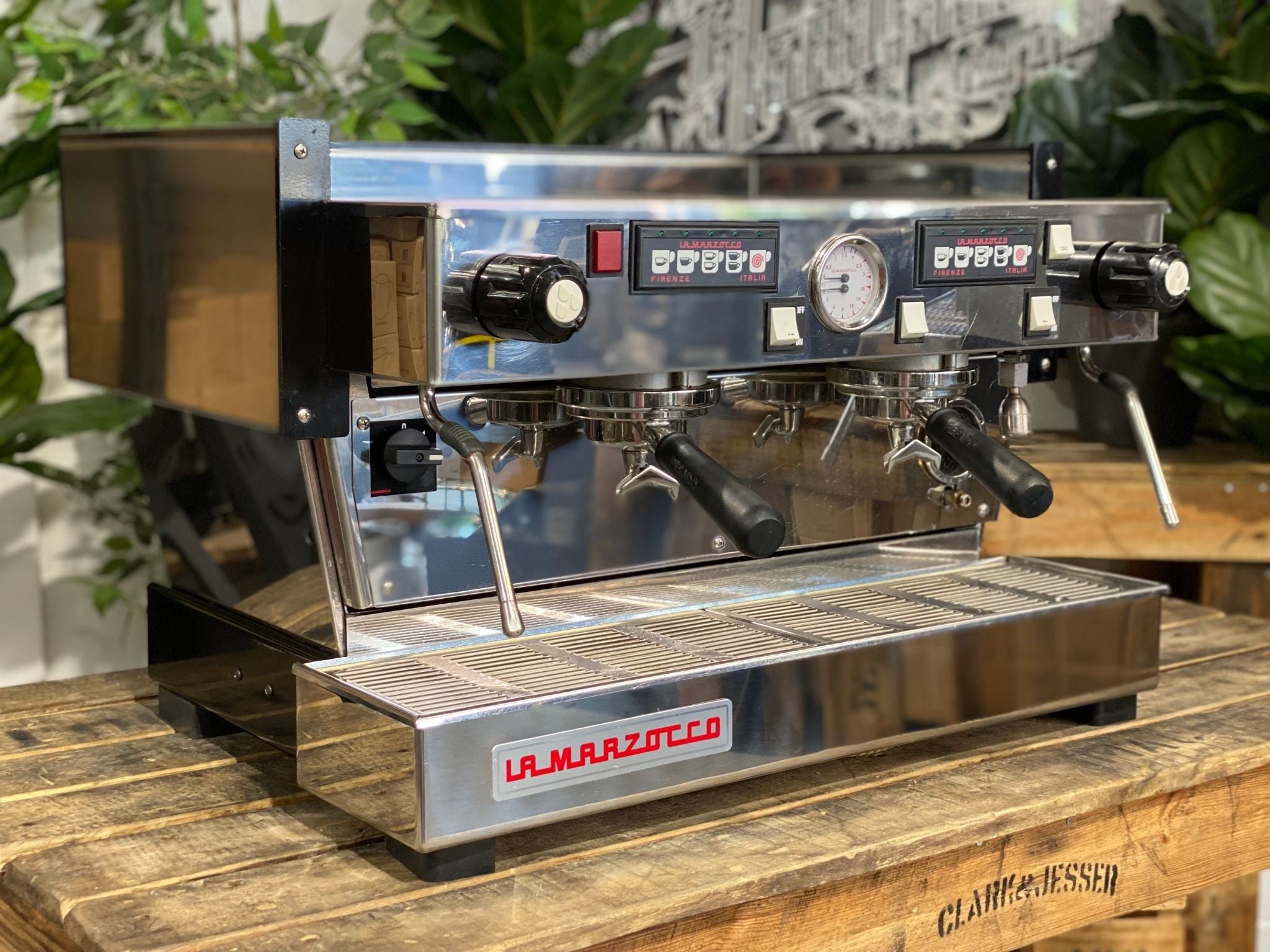 La Marzocco Linea Classic 2 Group Espresso Coffee Machine in Stainless steel is sitting on a wooden bench. There is greenery in the background. The image is taken from the front of the machine and the machine is sitting on an angle so that you can see the front of the machine and the side