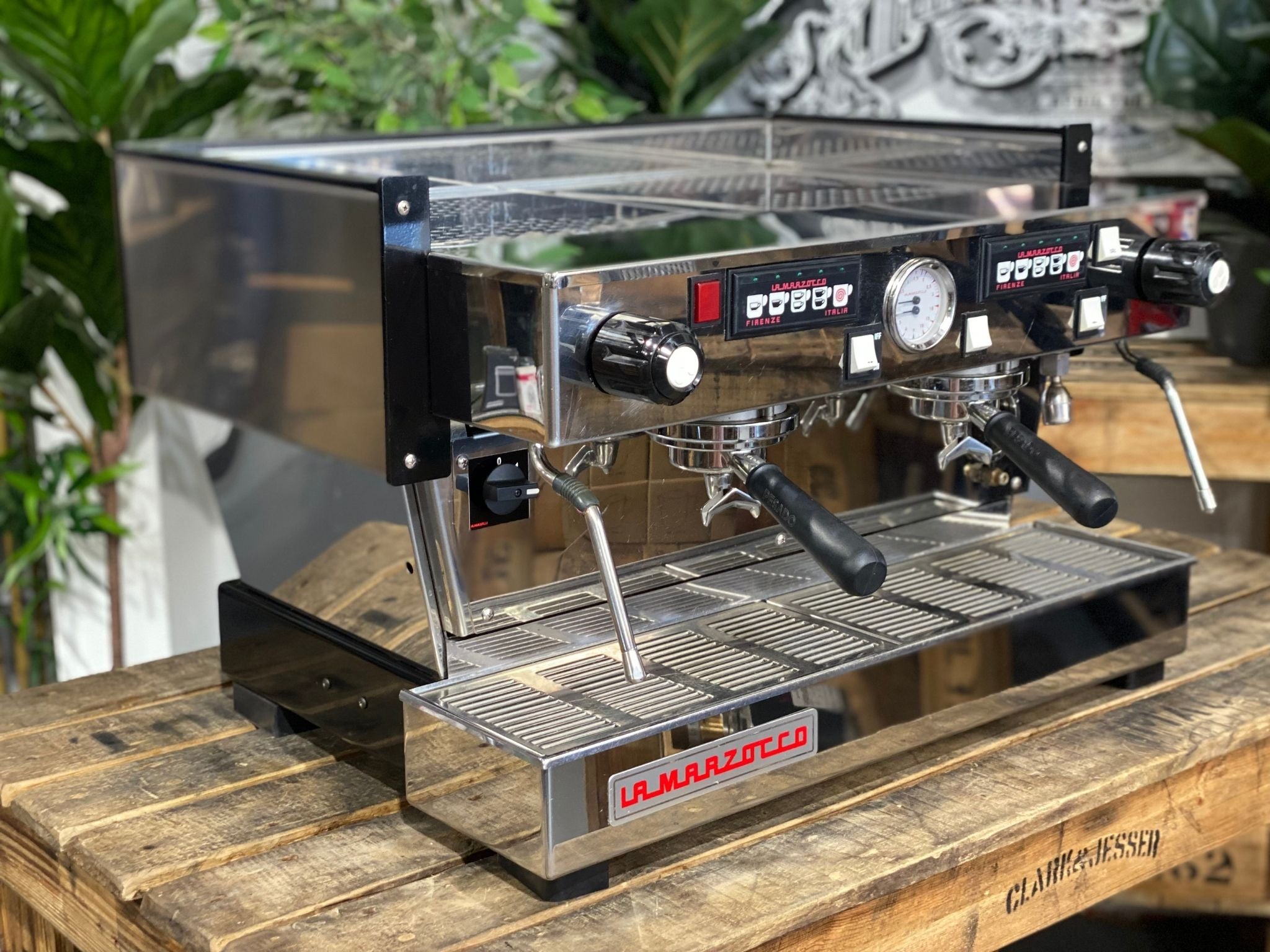 La Marzocco Linea Classic 2 Group Espresso Coffee Machine in Stainless steel is sitting on a wooden bench. There is greenery in the background. The image is taken from the front of the machine and the machine is sitting on an angle so that you can see the front of the machine and the side