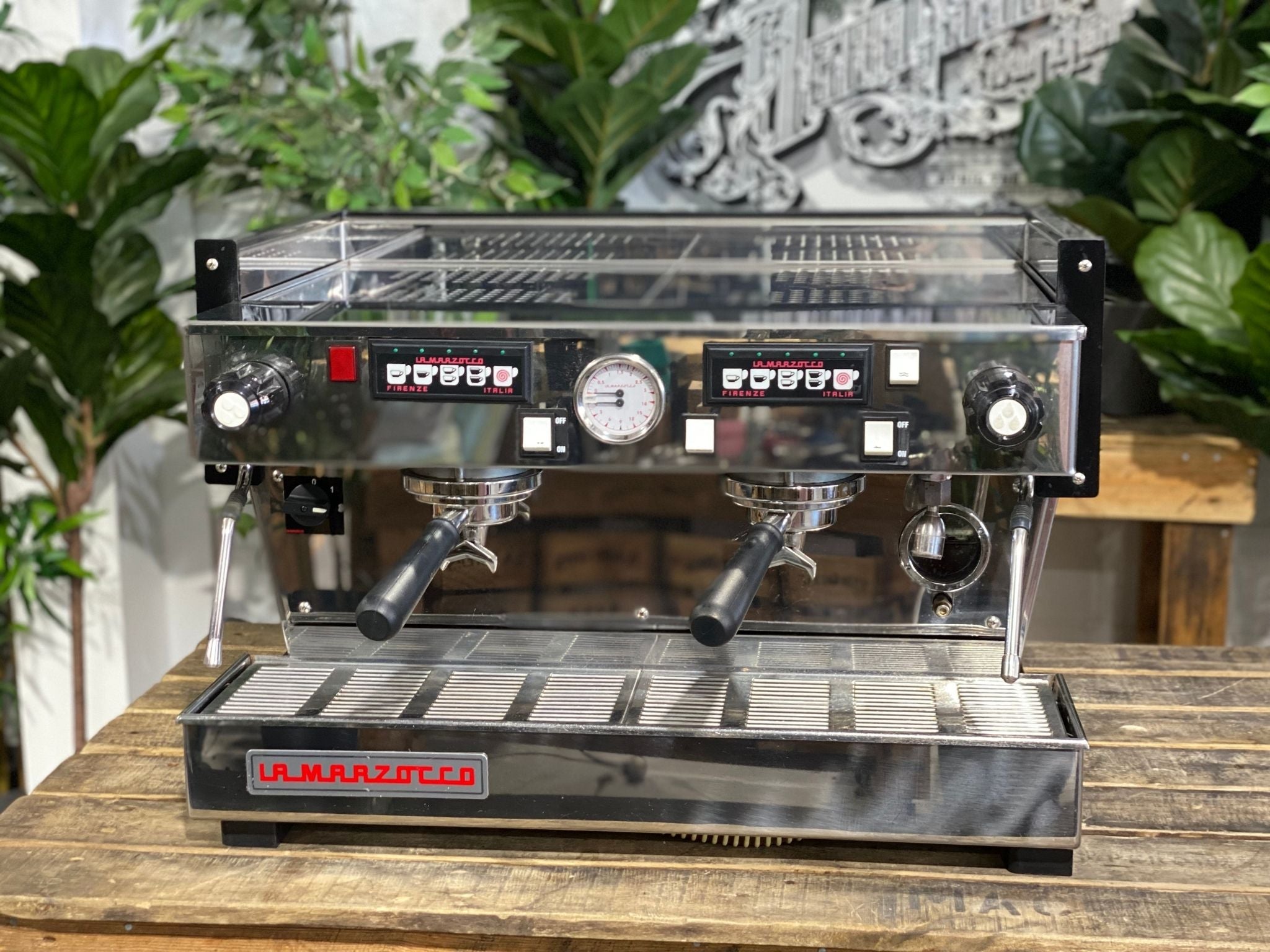 La Marzocco Linea Classic 2 Group Espresso Coffee Machine in Stainless steel sitting on a wooden bench. There is greenery in the background. The image is taken from the front of the machine so that you can see the front of the machine, control panel, buttons and group heads