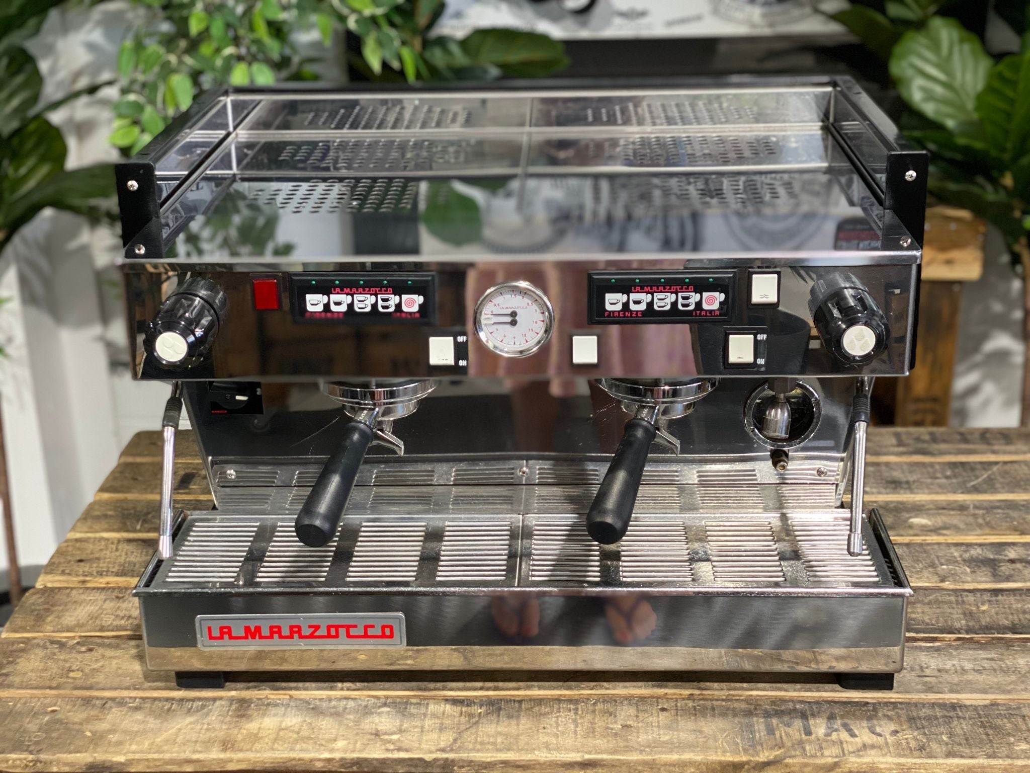 La Marzocco Linea Classic 2 Group Espresso Coffee Machine in Stainless steel sitting on a wooden bench. There is greenery in the background. The image is taken from the front of the machine but a higher angle so that you can see the front of the machine and also the top of the machine
