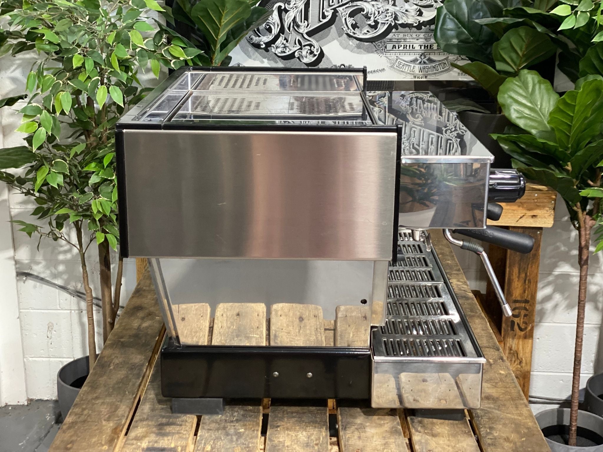 La Marzocco Linea Classic 2 Group Espresso Coffee Machine in Stainless steel sitting on a wooden bench. There is greenery in the background. The image is taken from the side of the machine.