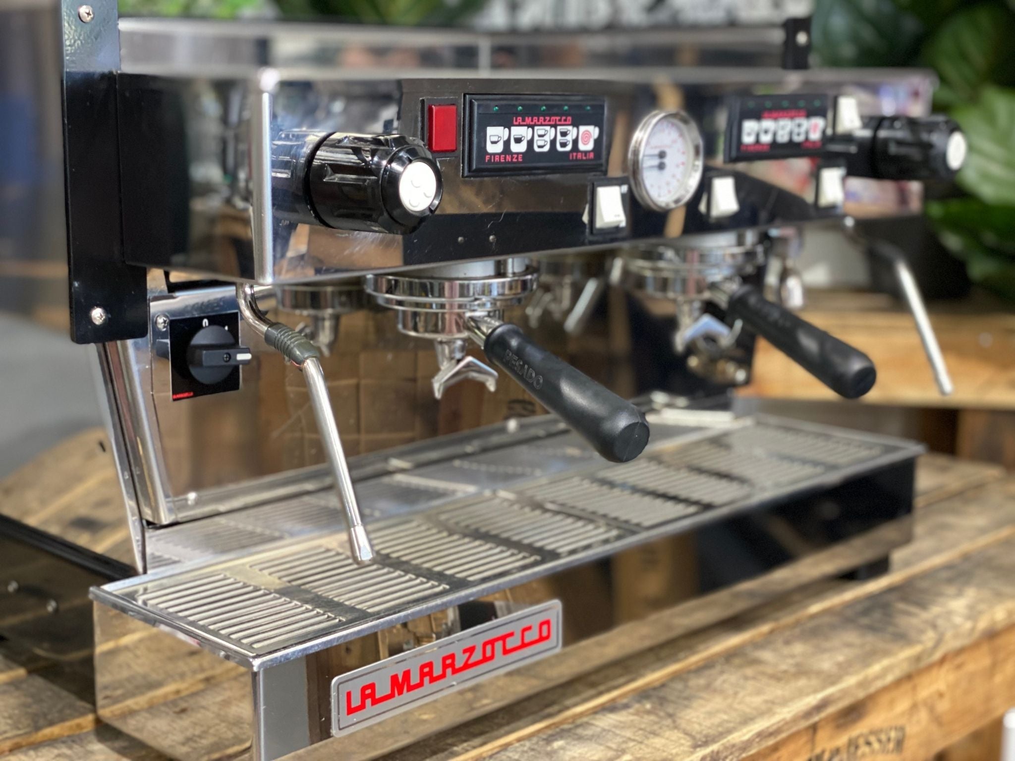 La Marzocco Linea Classic 2 Group Espresso Coffee Machine in Stainless steel is sitting on a wooden bench. There is greenery in the background. The image is a close up image taken from the front of the machine and the machine is sitting on an angle so that you can see the front of the machine, the controls including buttons, group handles and logo