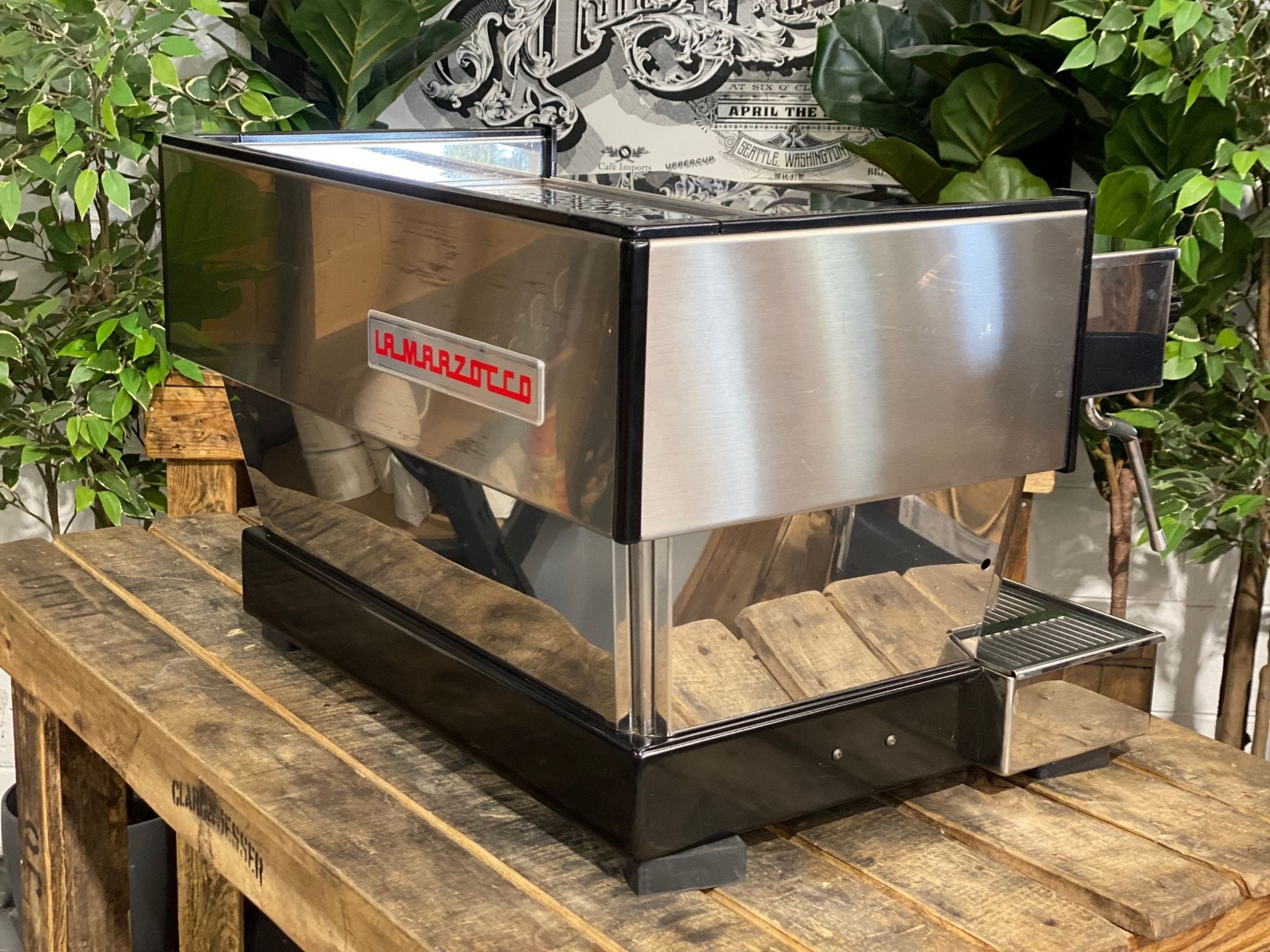 La Marzocco Linea Classic 2 Group Espresso Coffee Machine in Stainless steel sitting on a wooden bench. There is greenery in the background. The image is taken from behind the machine and the machine is sitting on an angle so that you can see the back of the machine and the side