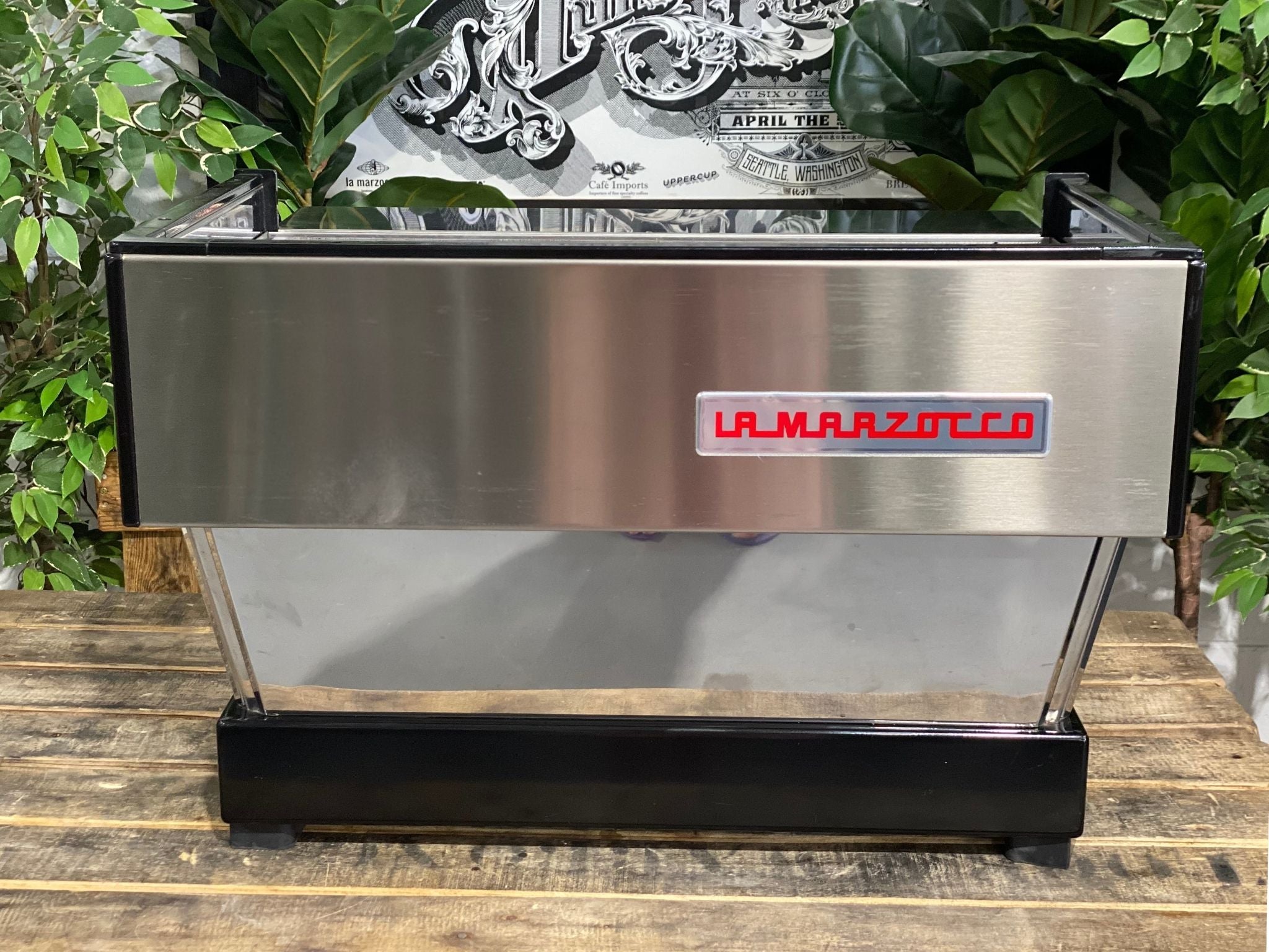 La Marzocco Linea Classic 2 Group Espresso Coffee Machine in Stainless steel is sitting on a wooden bench. There is greenery in the background. The image is taken from behind the machine so the back is visible. The back of the machine is a stainless steel panel with the red la marzocco logo