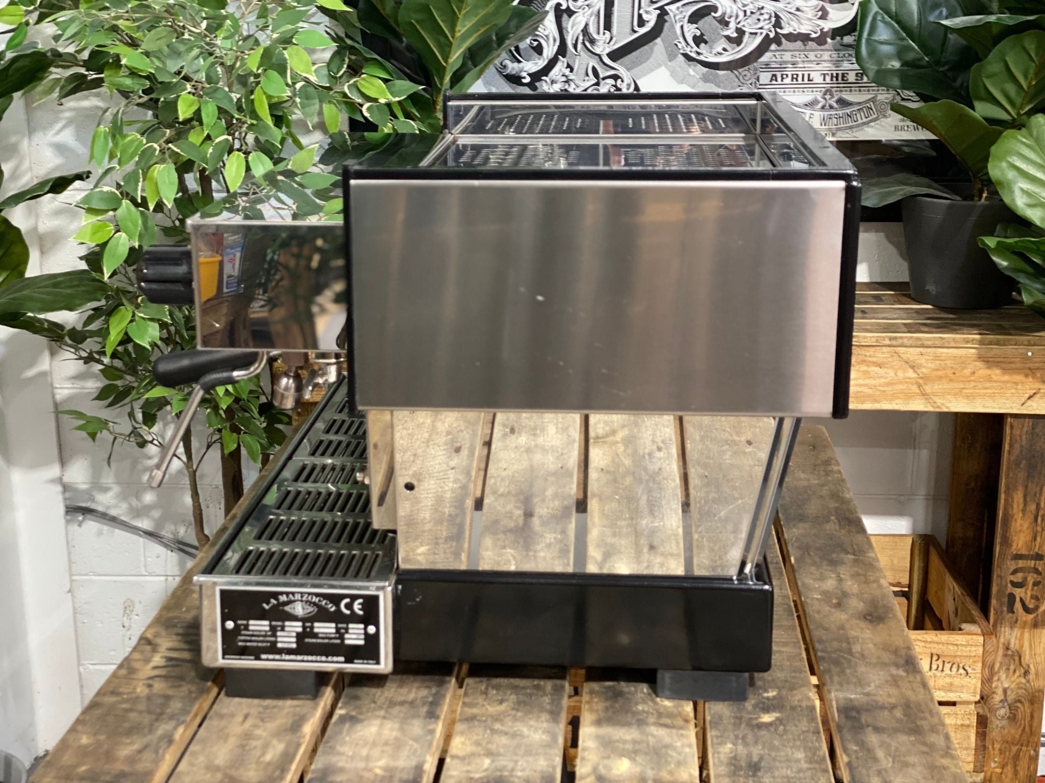 La Marzocco Linea Classic 2 Group Espresso Coffee Machine in Stainless steel sitting on a wooden bench. There is greenery in the background. The image is taken from the side of the machine.