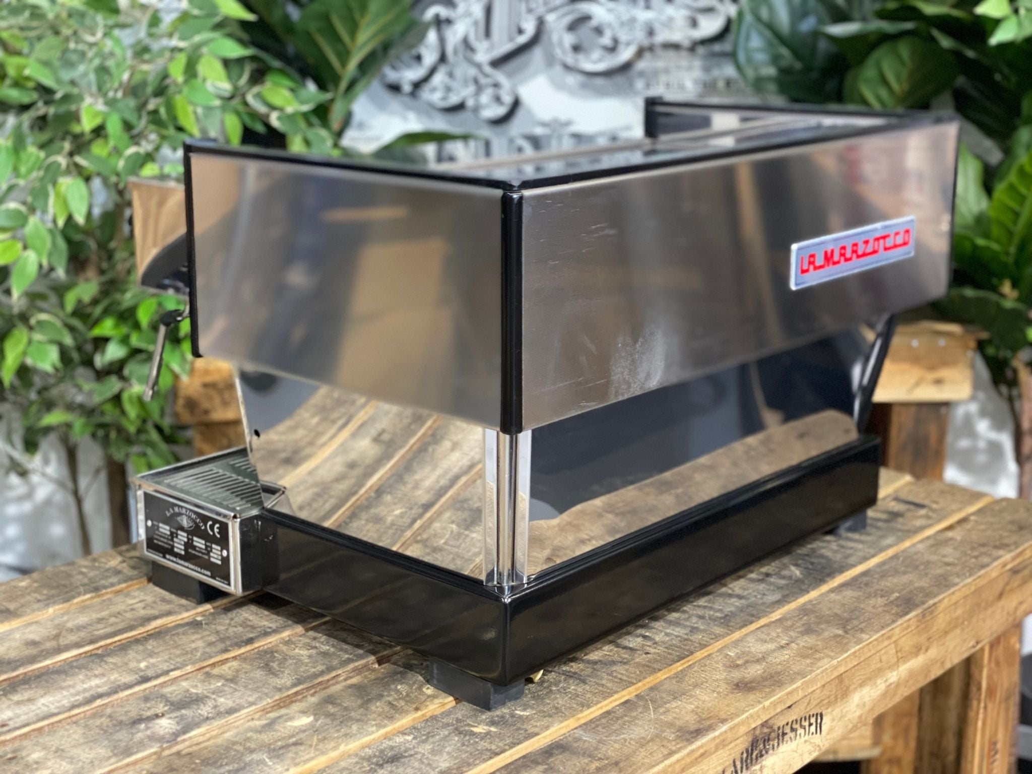 La Marzocco Linea Classic 2 Group Espresso Coffee Machine in Stainless steel sitting on a wooden bench. There is greenery in the background. The image is taken from behind the machine and the machine is sitting on an angle so that you can see the back of the machine and the side