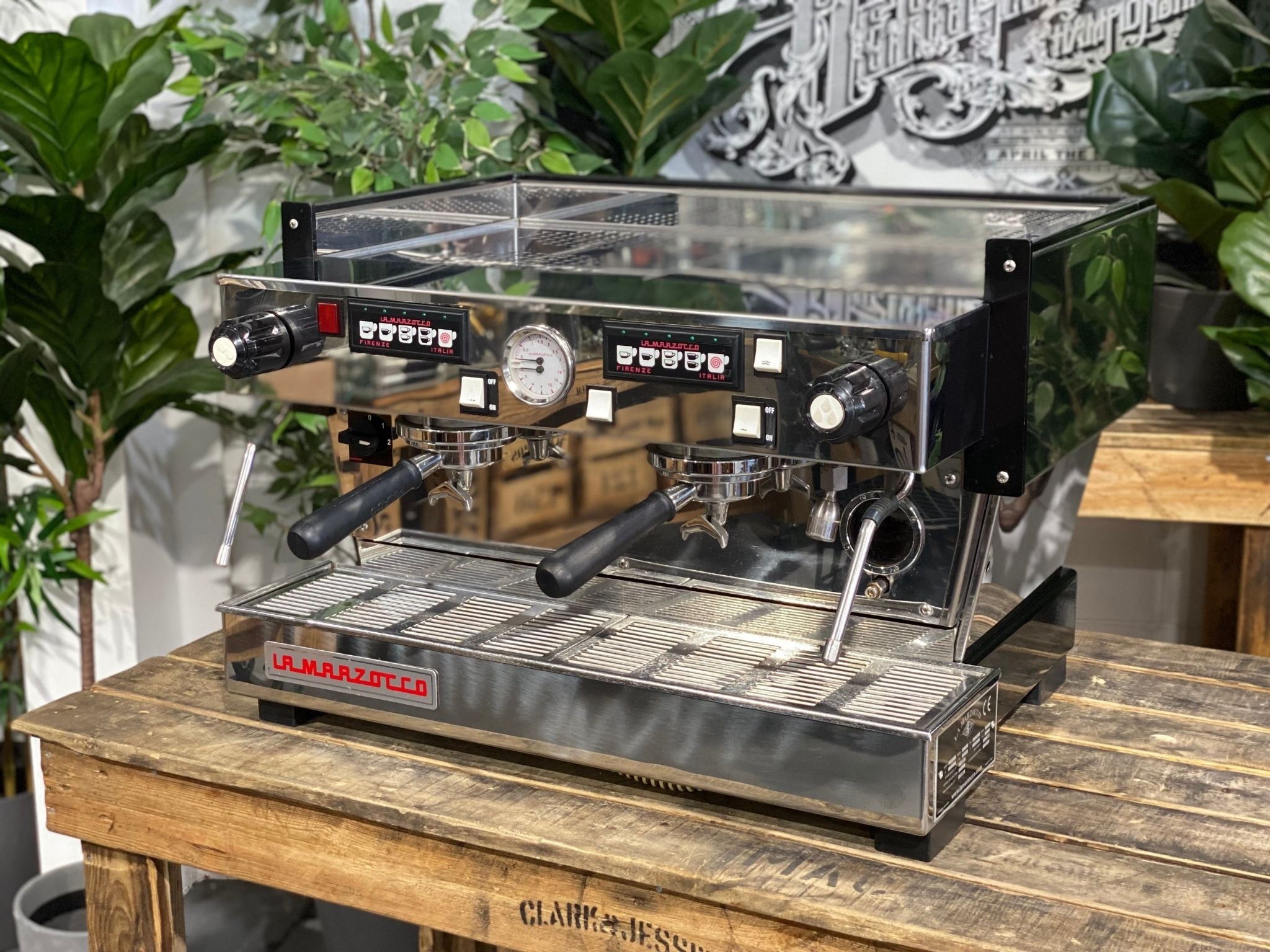 La Marzocco Linea Classic 2 Group Espresso Coffee Machine in Stainless steel is sitting on a wooden bench. There is greenery in the background. The image is taken from the front of the machine and the machine is sitting on an angle so that you can see the front of the machine and the side