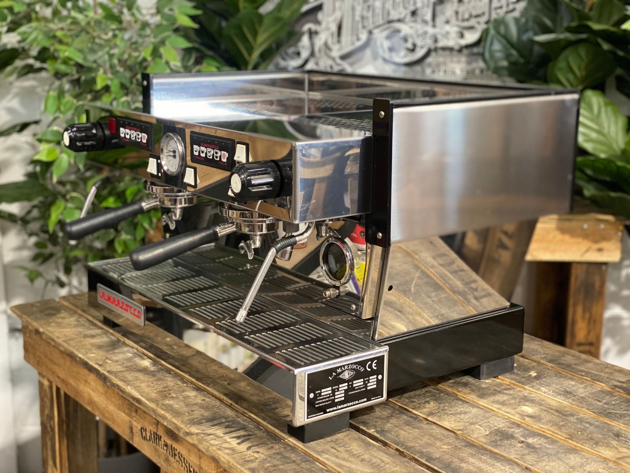 La Marzocco Linea Classic 2 Group Espresso Coffee Machine in Stainless steel sitting on a wooden bench. There is greenery in the background. The image is taken from the side of the machine.
