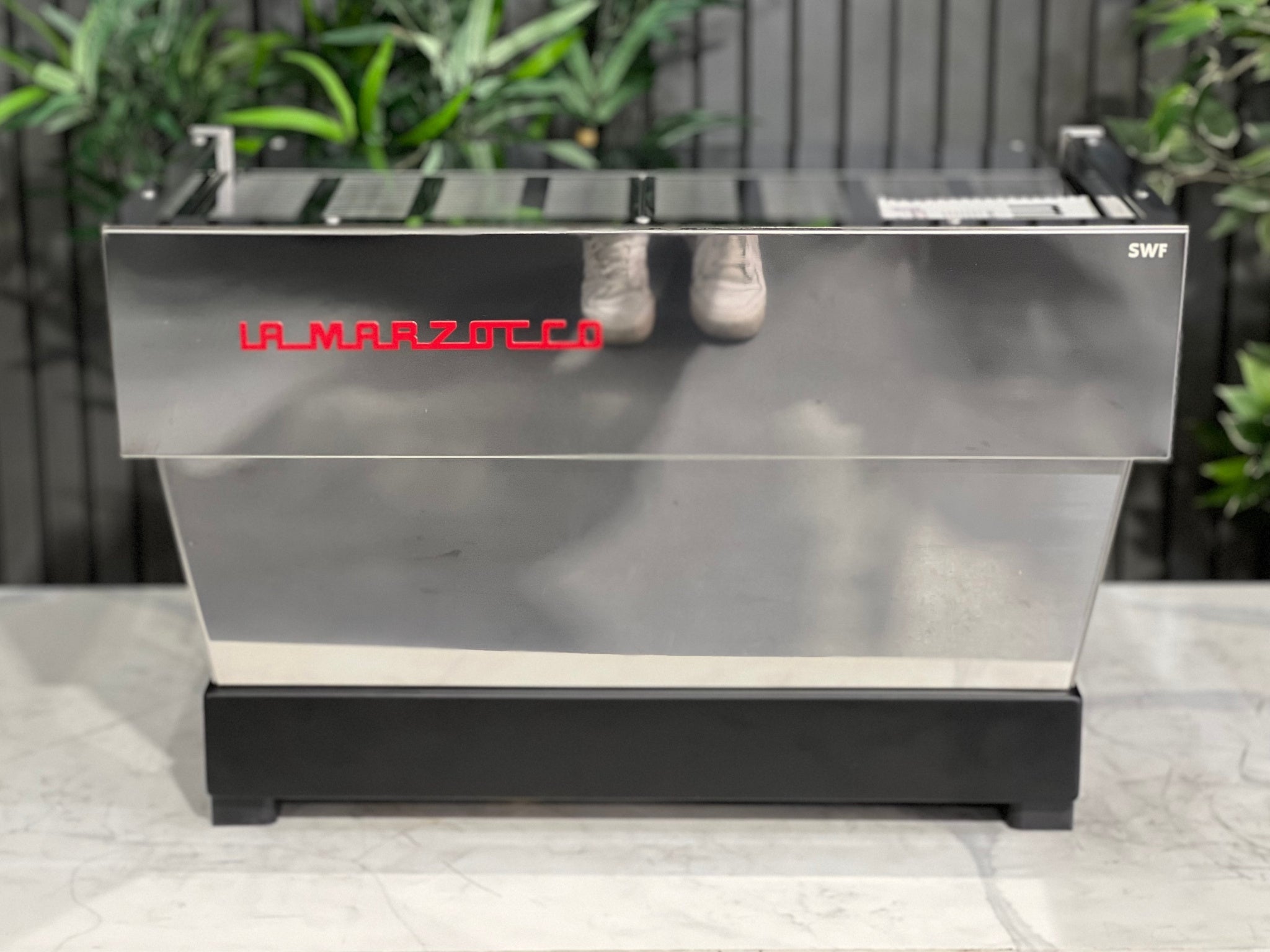 A New La Marzocco Linea Classic S 2 Group in Stainless steel  is sitting on a marble bench. There is greenery in the background. The image is taken from behind the machine so the back is visible. The back of the machine is a stainless steel panel with the red la marzocco logo