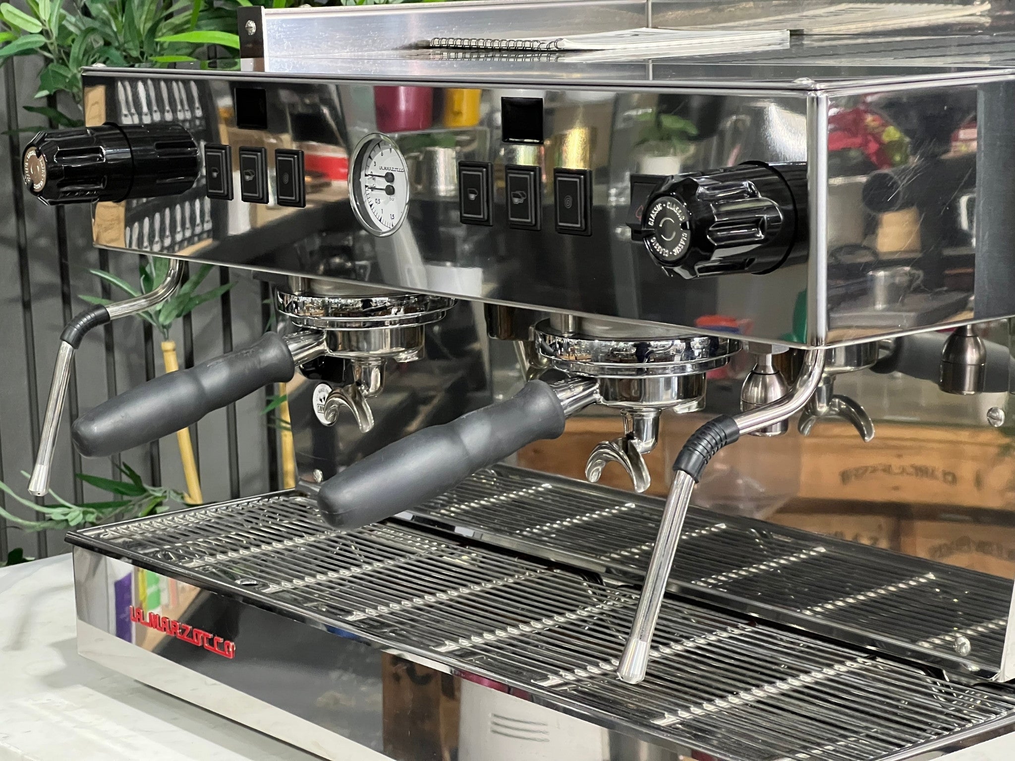 A New La Marzocco Linea Classic S 2 Group in Stainless steel  is sitting on a marble bench. There is greenery in the background. The image is a close up image taken from the front of the machine and the machine is sitting on an angle so that you can see the front of the machine, the controls including buttons, group handles and logo
