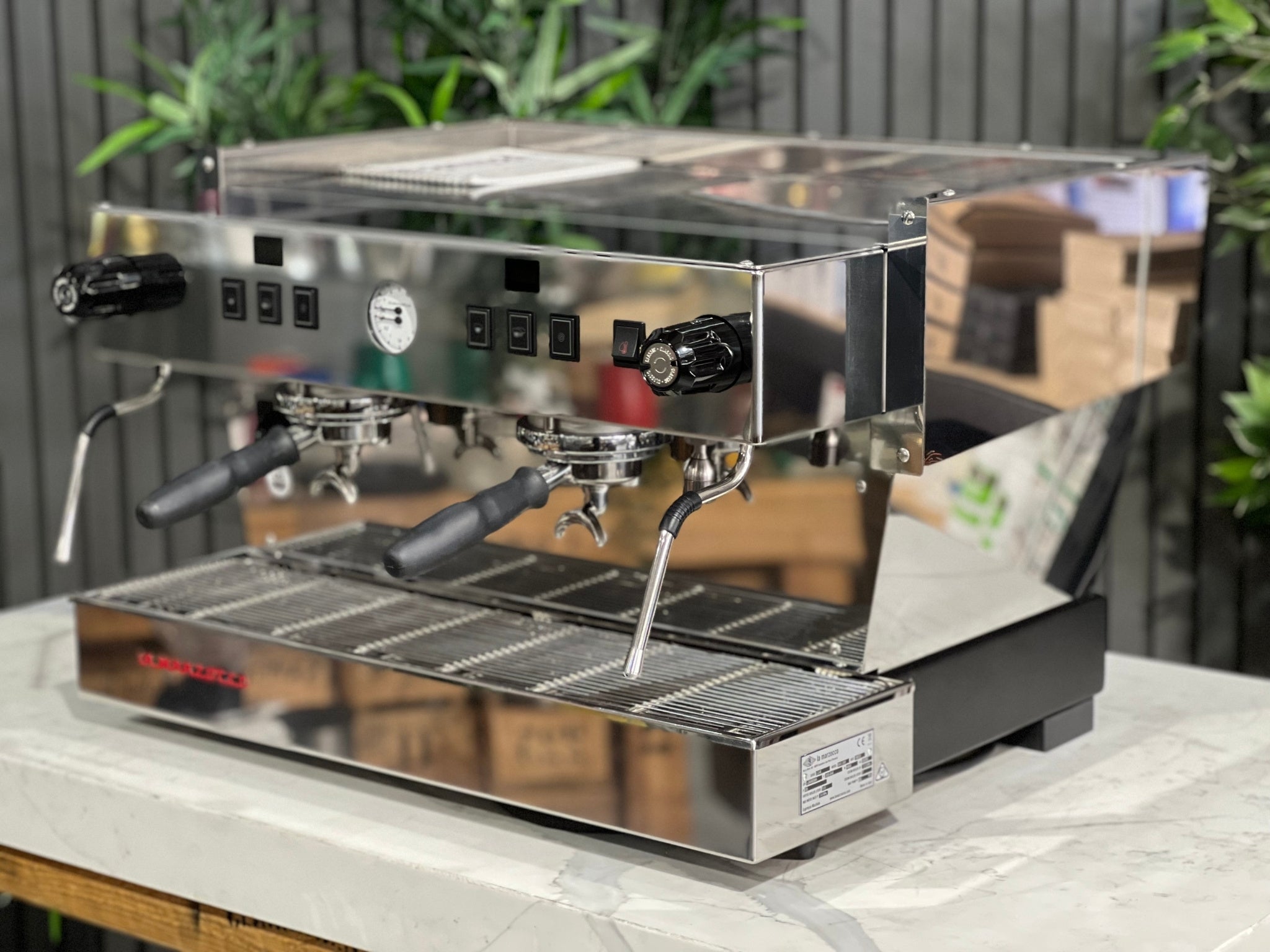 La Marzocco Linea Classic S AV 2 Group Coffee Machine Stainless - New showing an angled view, highlighting the dual steam wands, group heads, and stainless steel body, placed on a marble bench with greenery in the background for a premium café setting.