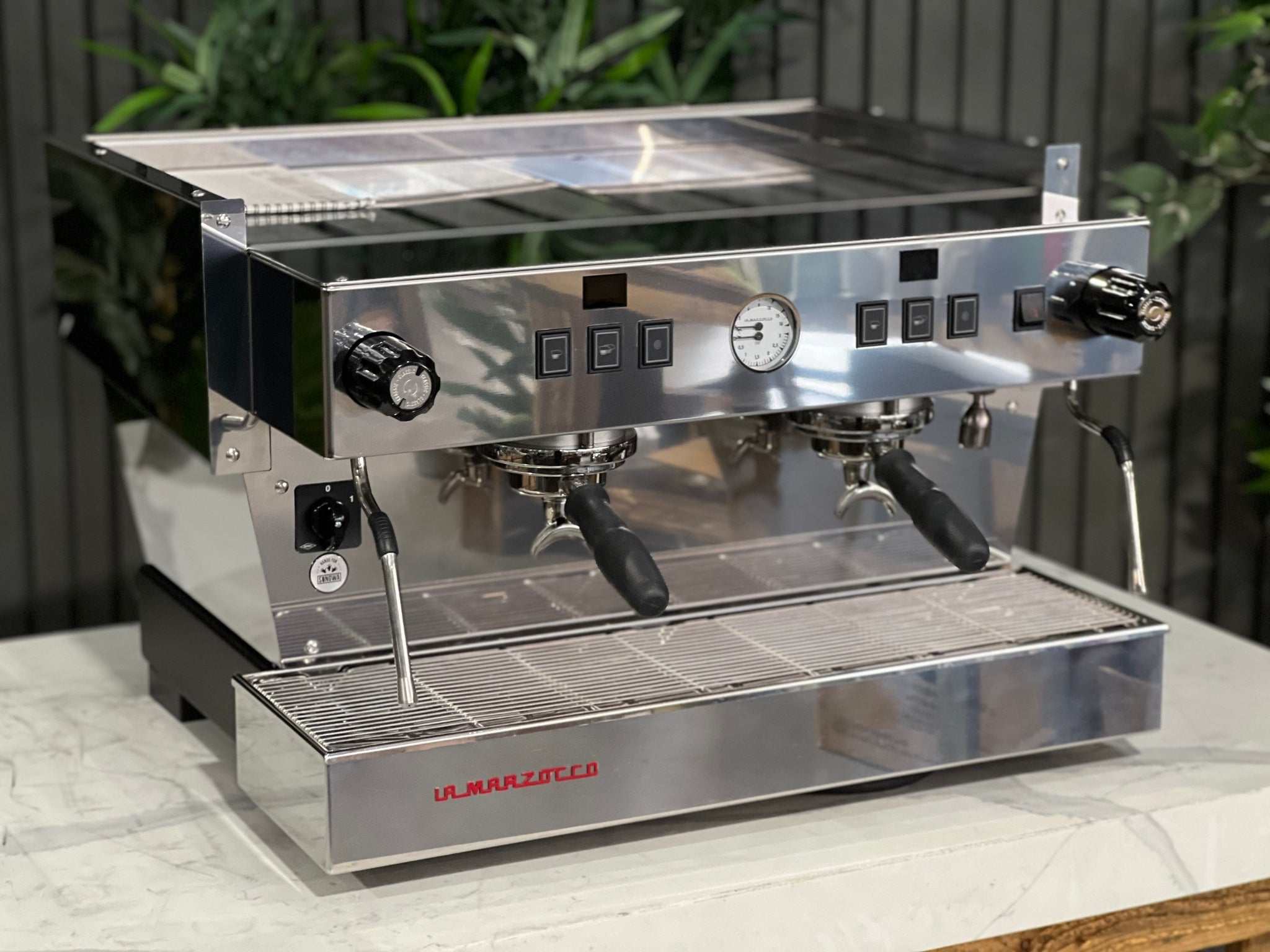 A New La Marzocco Linea Classic S 2 Group in Stainless steel is sitting on a marble bench. There is greenery in the background. The image is taken from the front of the machine and the machine is sitting on an angle so that you can see the front of the machine and the side