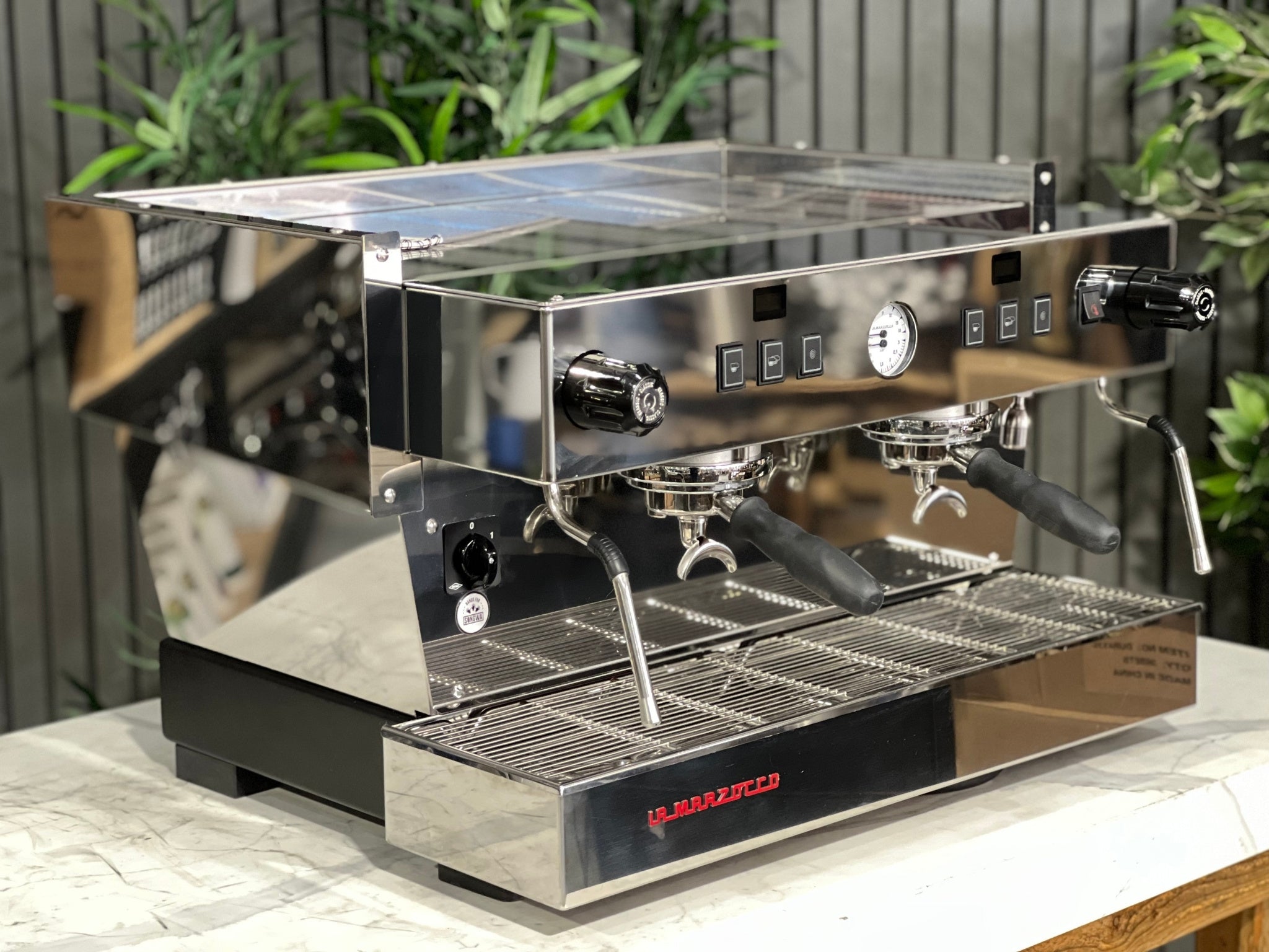 A New La Marzocco Linea Classic S 2 Group in Stainless steel  is sitting on a marble bench. There is greenery in the background. The image is taken from the front of the machine and the machine is sitting on an angle so that you can see the front of the machine and the side