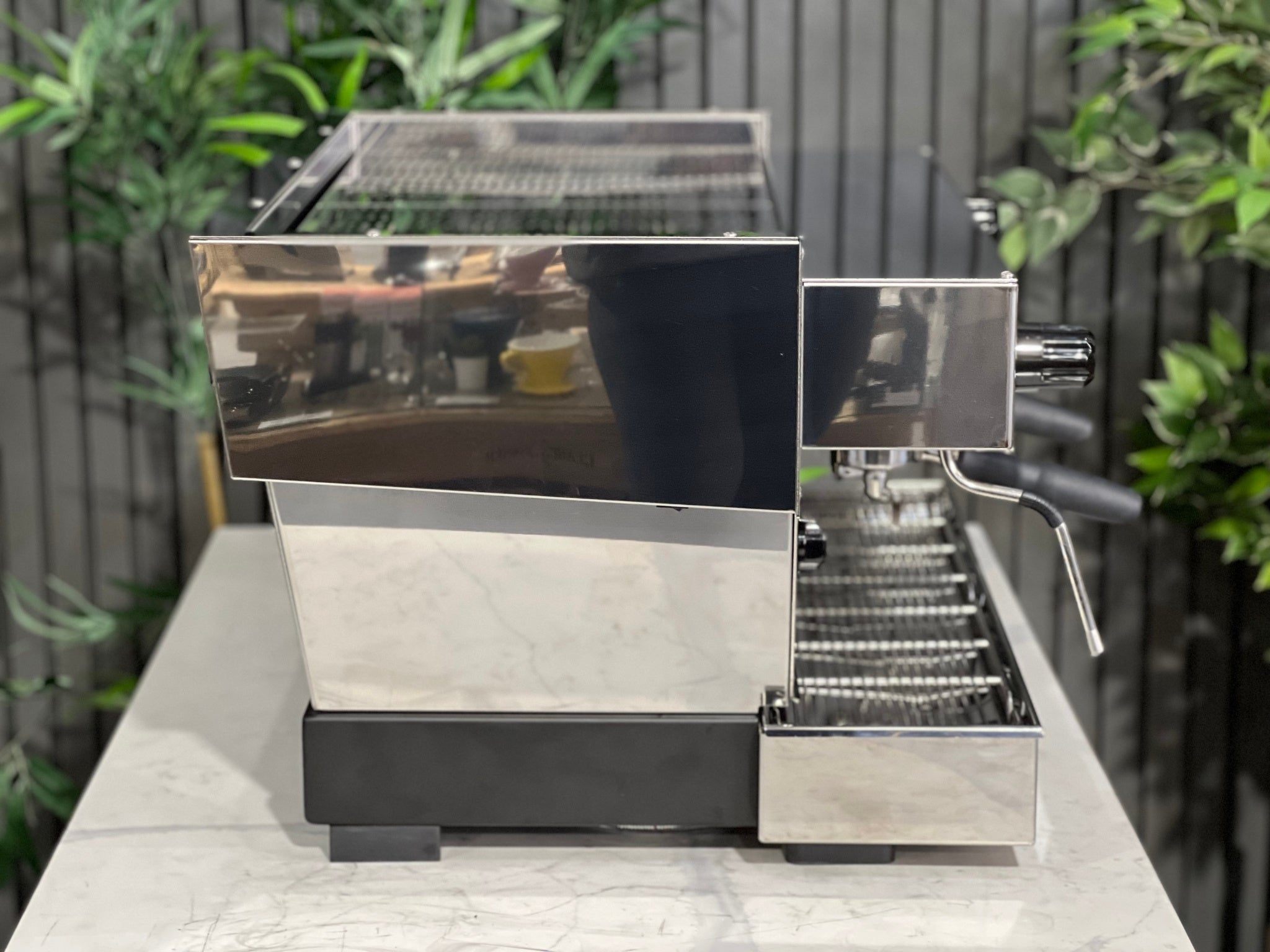 A New La Marzocco Linea Classic S 2 Group in Stainless steel sitting on a marble bench. There is greenery in the background. The image is taken from the side of the machine.