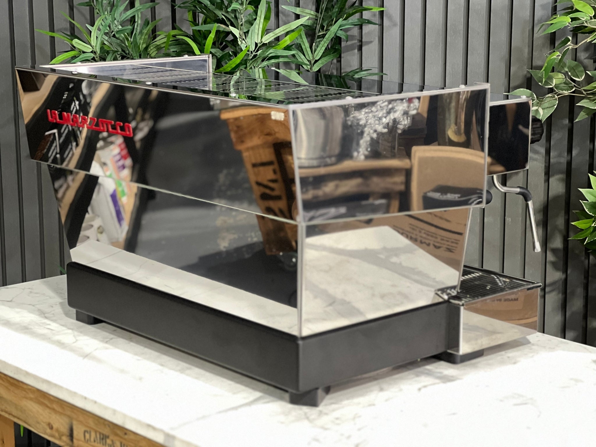 A New La Marzocco Linea Classic S 2 Group in Stainless steel  sitting on a marble bench. There is greenery in the background. The image is taken from behind the machine and the machine is sitting on an angle so that you can see the back of the machine and the side