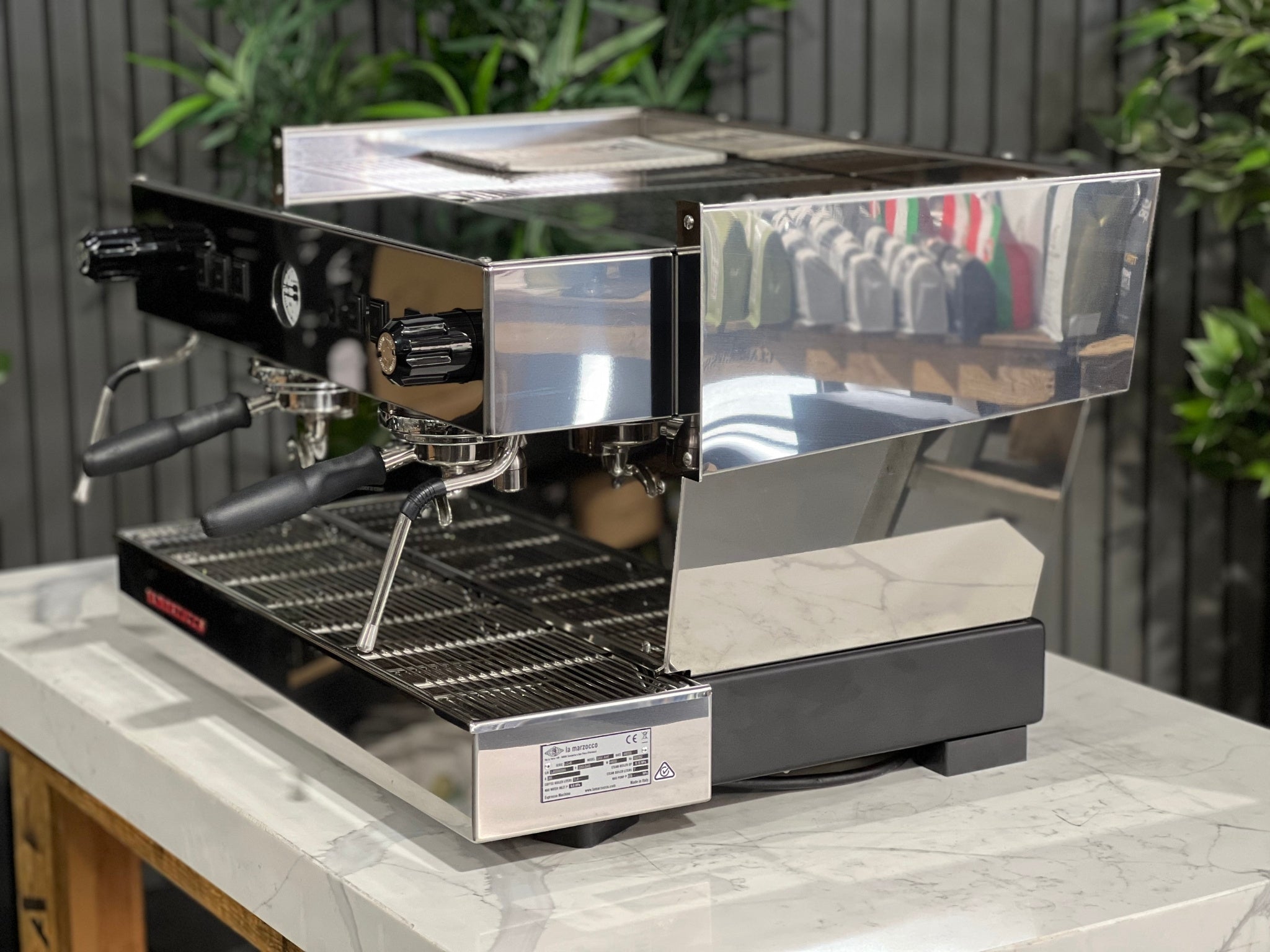 A New La Marzocco Linea Classic S 2 Group in Stainless steel is sitting on a marble bench. There is greenery in the background. The image is taken from the front of the machine and the machine is sitting on an angle so that you can see the front of the machine and the side