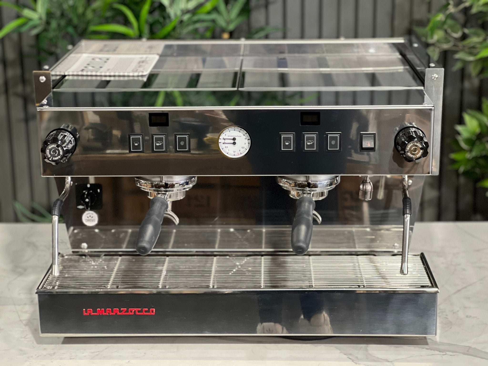 A New La Marzocco Linea Classic S 2 Group in Stainless steel  sitting on a marble bench. There is greenery in the background. The image is taken from the front of the machine but a higher angle so that you can see the front of the machine and also the top of the machine