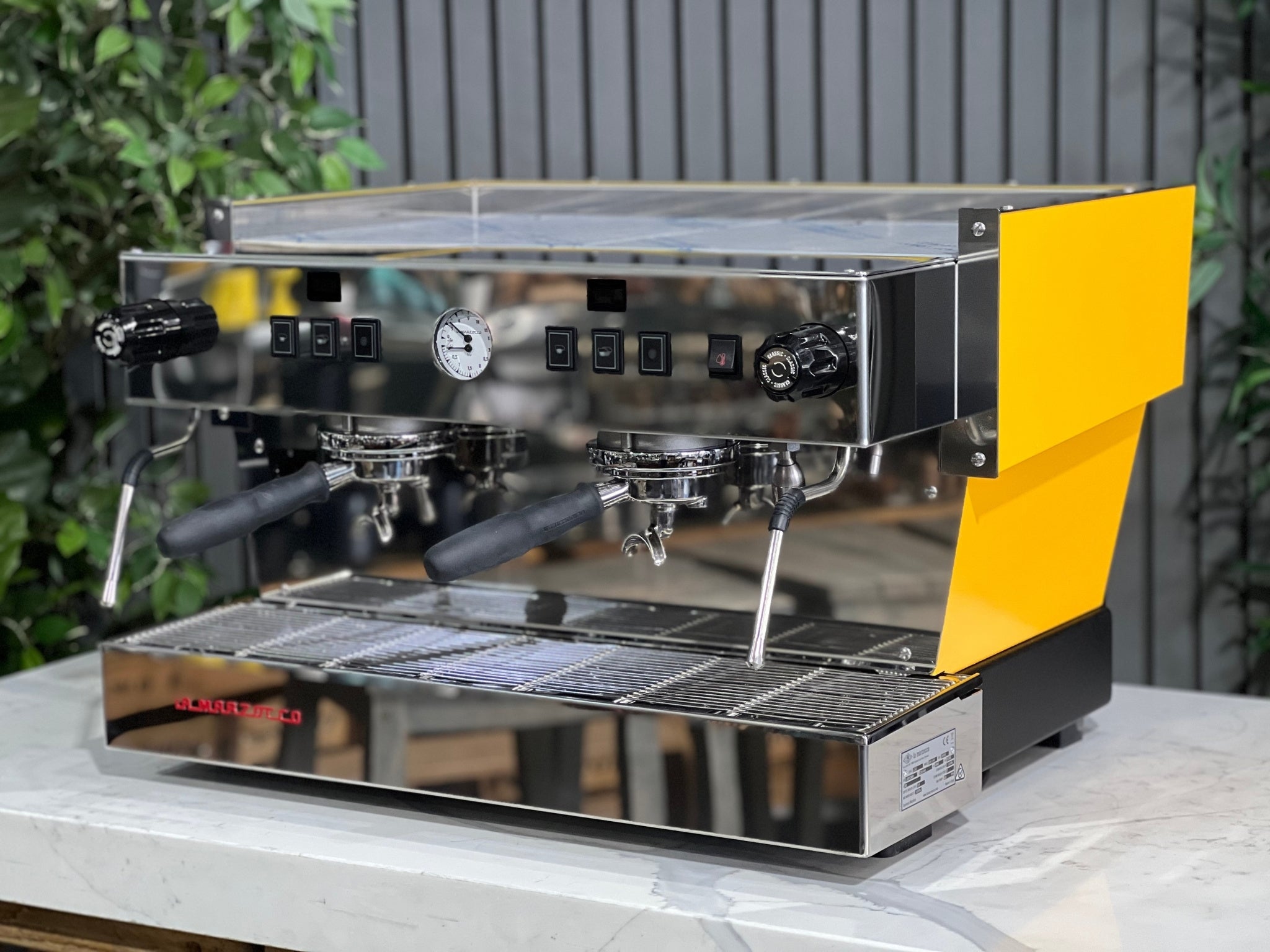 A new La Marzocco Linea Classic S AV 2 Group Coffee Machine in Yellow is sitting on a marble bench. There is greenery in the background. The image is taken from the front of the machine and the machine is sitting on an angle so that you can see the front of the machine and the side.