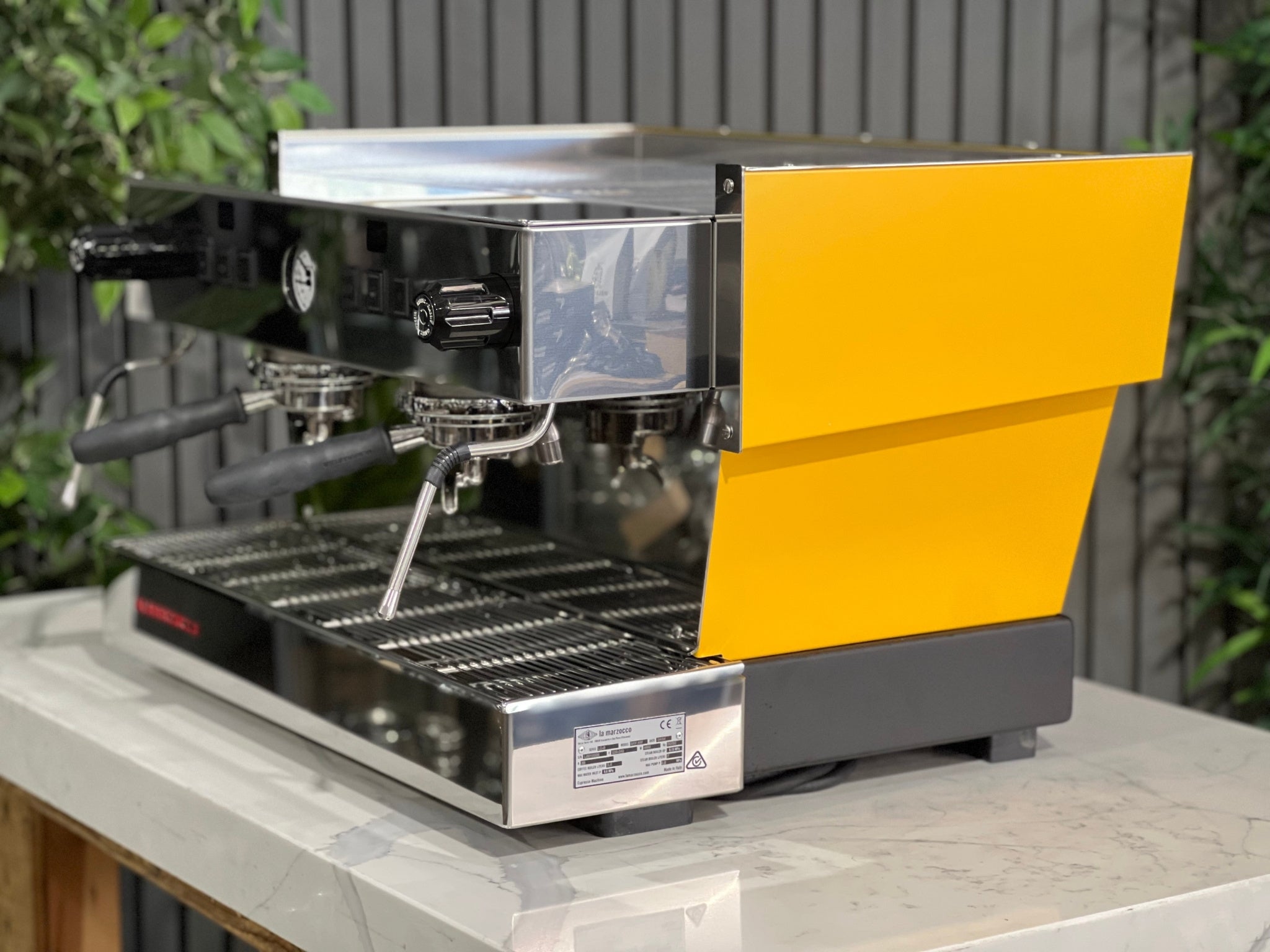 A new La Marzocco Linea Classic S AV 2 Group Coffee Machine in Yellow  sitting on a marble bench. There is greenery in the background. The image is taken from the side of the machine.