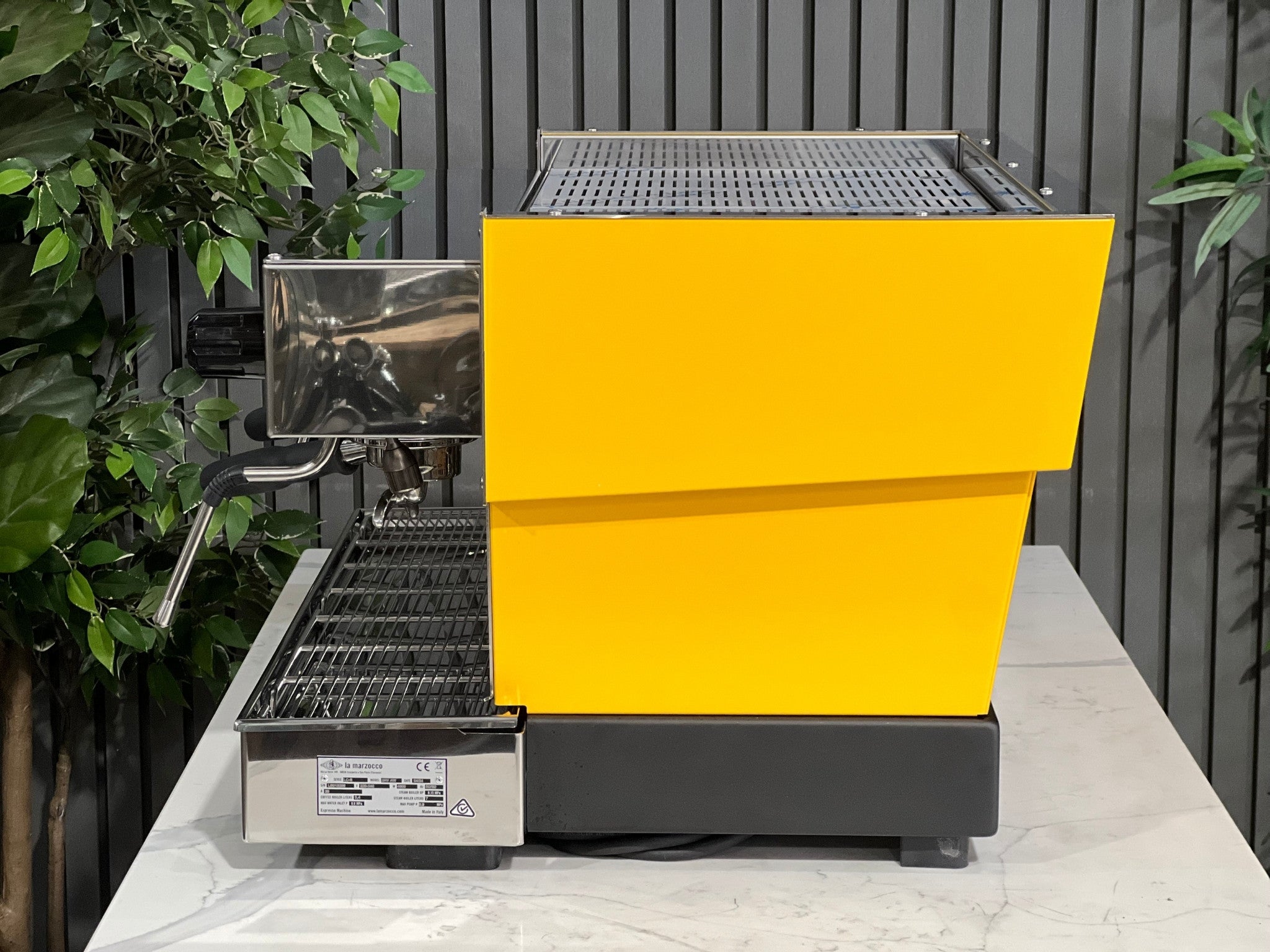 A new La Marzocco Linea Classic S AV 2 Group Coffee Machine in Yellow  sitting on a marble bench. There is greenery in the background. The image is taken from the side of the machine.