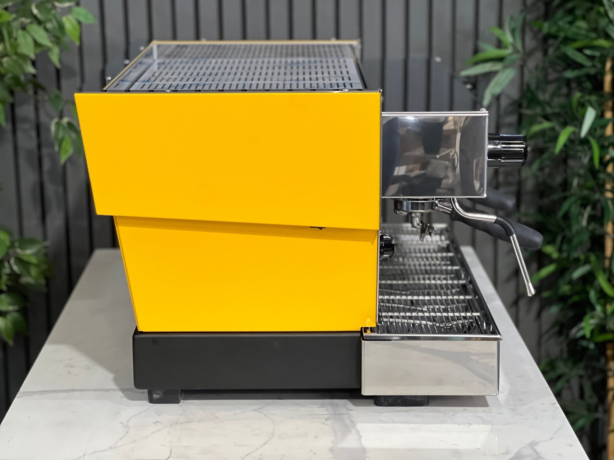 A new La Marzocco Linea Classic S AV 2 Group Coffee Machine in Yellow  sitting on a marble bench. There is greenery in the background. The image is taken from the side of the machine.