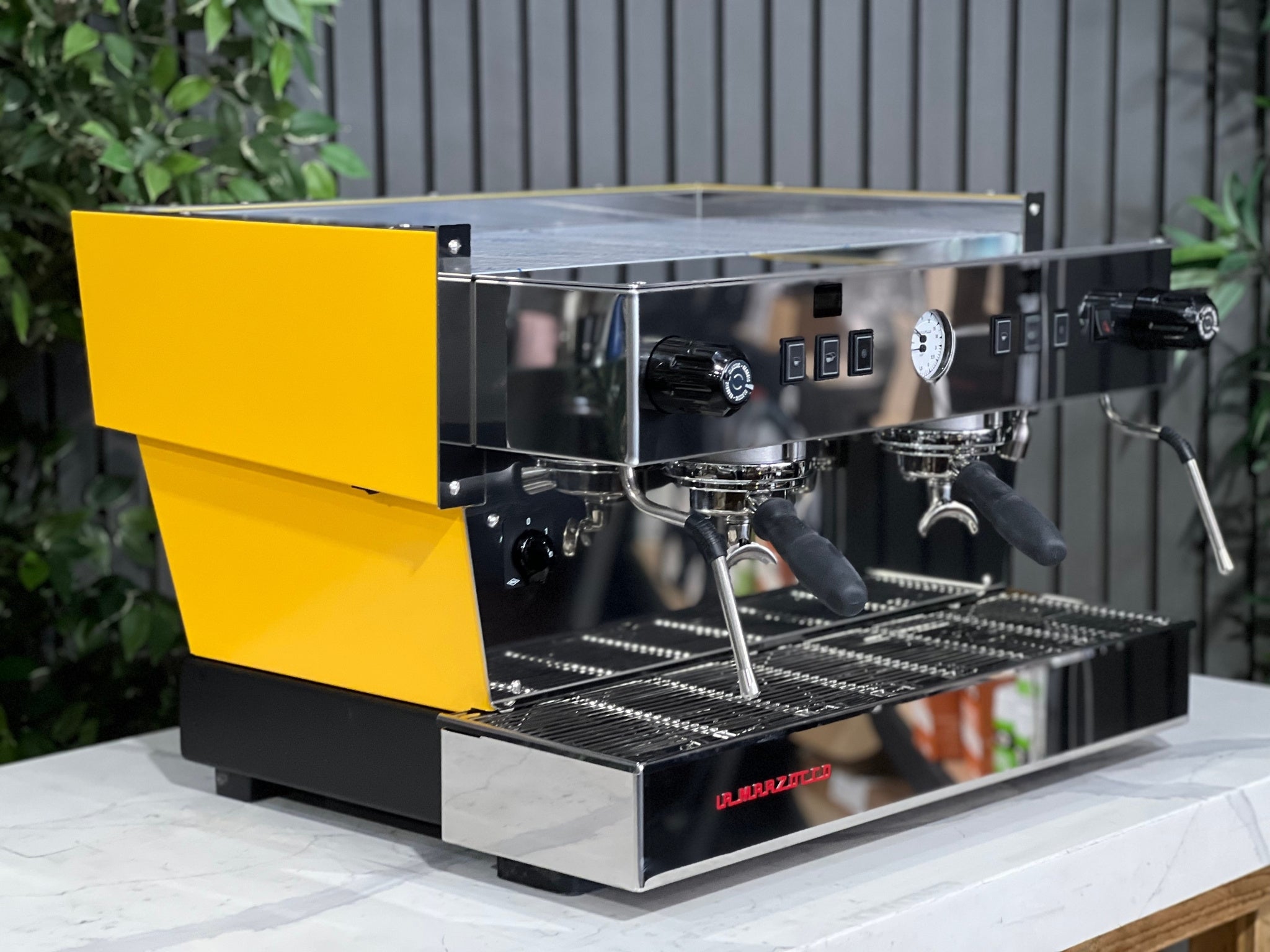 A new La Marzocco Linea Classic S AV 2 Group Coffee Machine in Yellow  is sitting on a marble bench. There is greenery in the background. The image is taken from the front of the machine and the machine is sitting on an angle so that you can see the front of the machine and the side