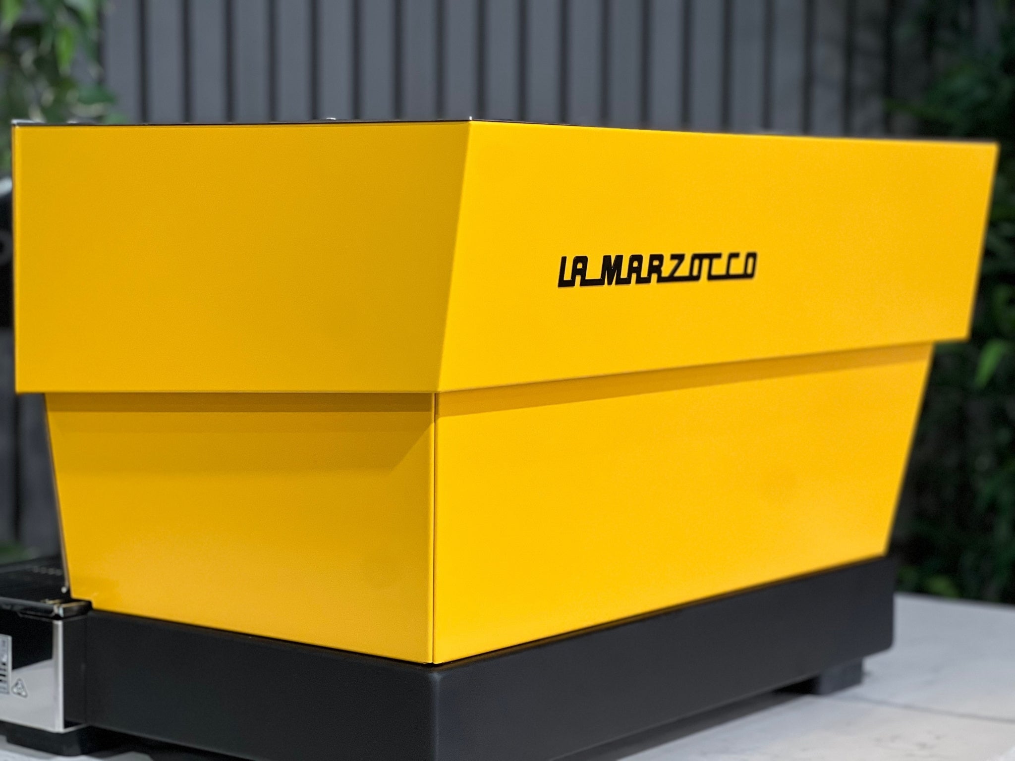 A new La Marzocco Linea Classic S AV 2 Group Coffee Machine in Yellow  is sitting on a marble bench. There is greenery in the background. The image is a close up image taken from the back of the machine and the machine is sitting on an angle, the canary yellow back panels and black la marzocco logo is visible.