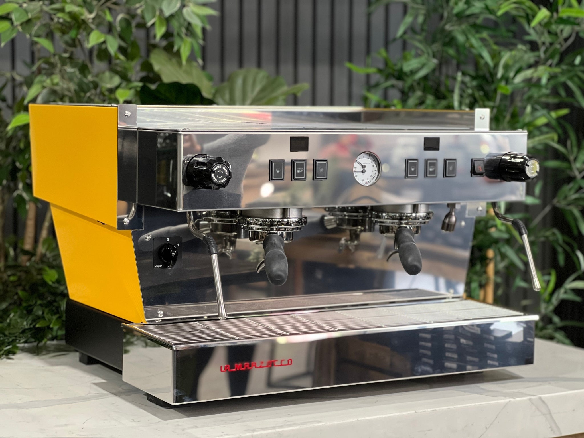 A new La Marzocco Linea Classic S AV 2 Group Coffee Machine in Yellow  is sitting on a marble bench. There is greenery in the background. The image is taken from the front of the machine and the machine is sitting on an angle so that you can see the front of the machine and the side .