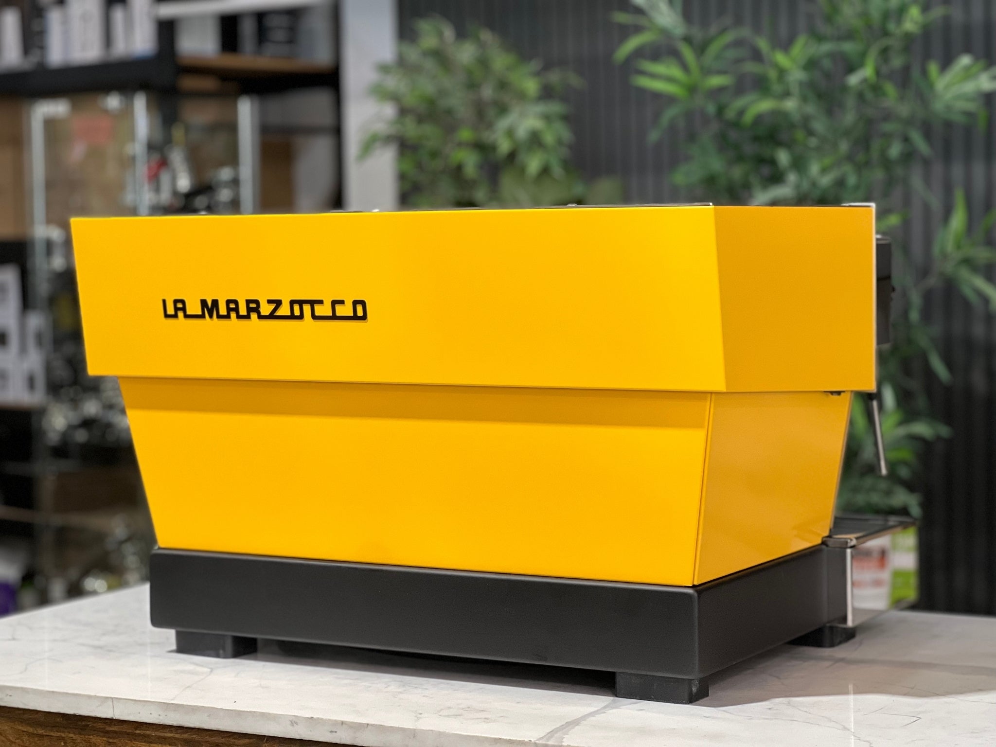 A new La Marzocco Linea Classic S AV 2 Group Coffee Machine in Yellow sitting on a marble bench. There is greenery in the background. The image is taken from behind the machine and the machine is sitting on an angle so that you can see the back of the machine and the side.