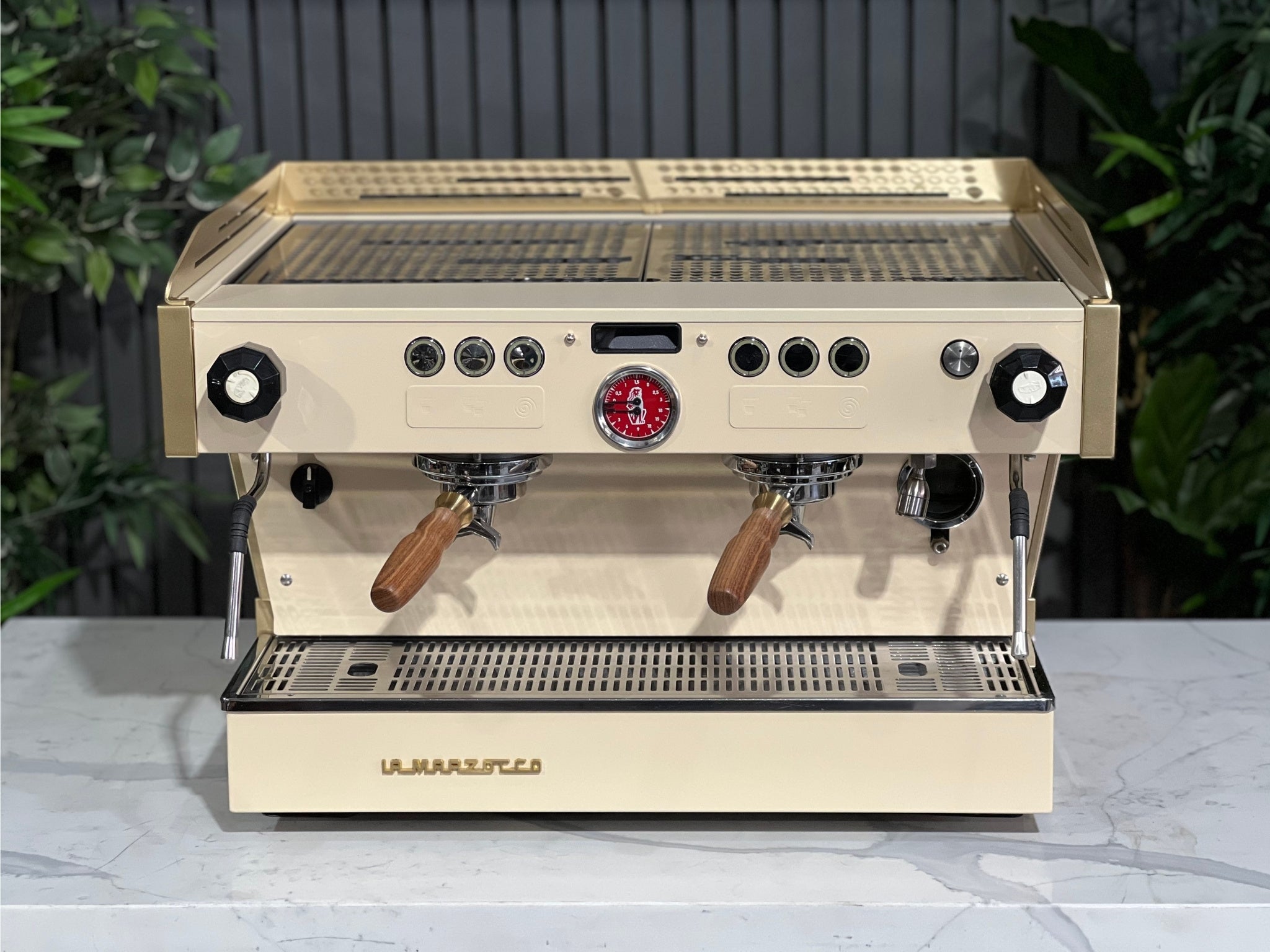 La Marzocco Linea PB 2 Group Coffee Machine Cream & Gold - Used seen from a higher front angle, with the cup tray and top of the espresso machine visible. Set on a marble bench with greenery behind, highlighting its sleek build and espresso-ready design.