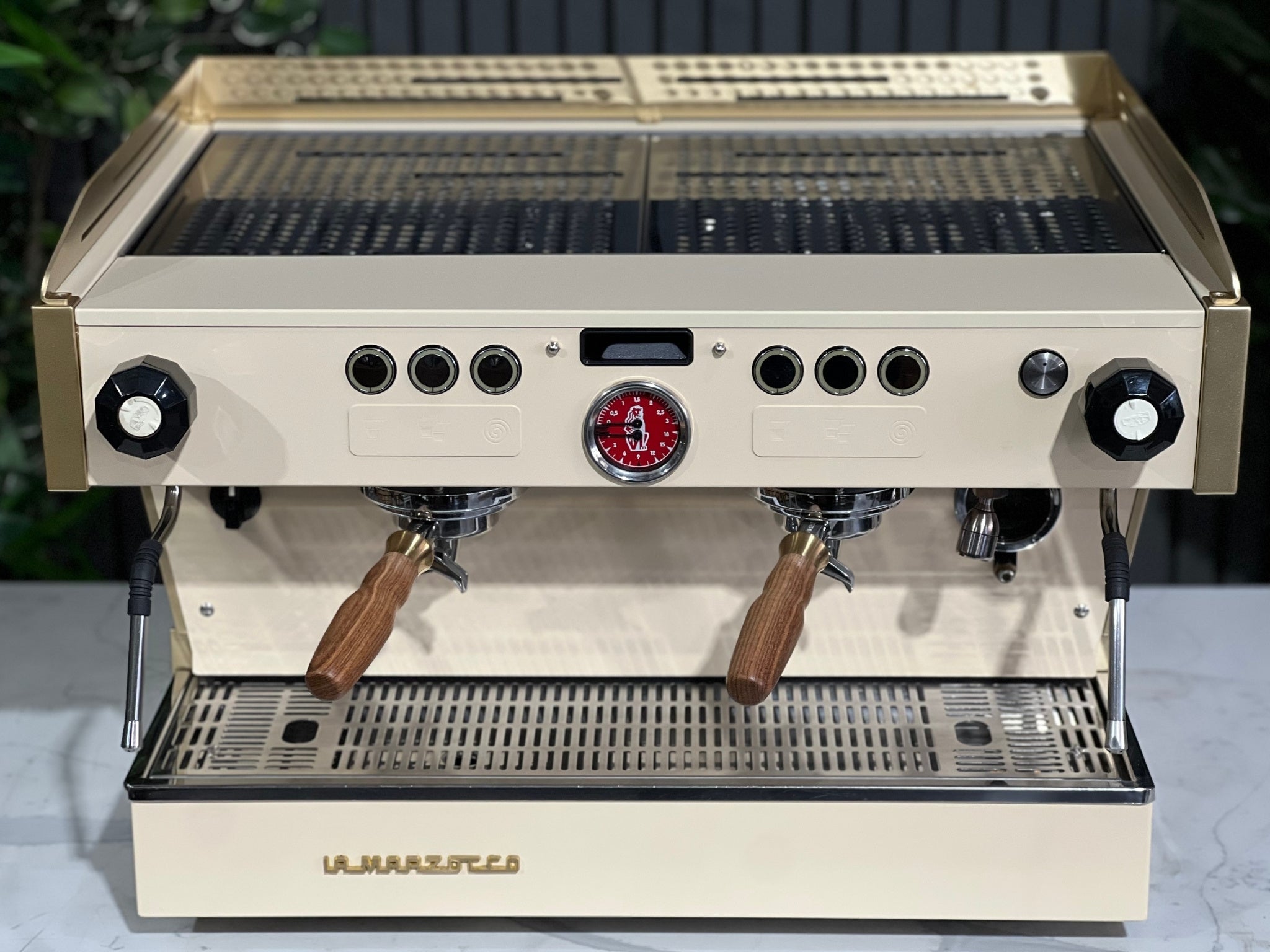 La Marzocco Linea PB 2 Group Coffee Machine Cream & Gold - Used seen from a higher front angle, with the cup tray and top of the espresso machine visible. Set on a marble bench with greenery behind, highlighting its sleek build and espresso-ready design.